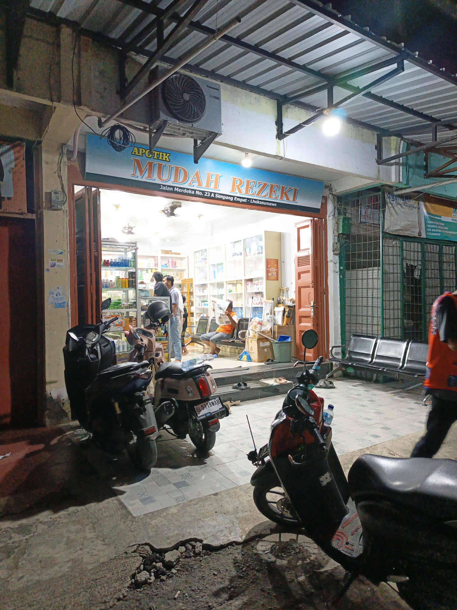 The image depicts the exterior of a pharmacy named "Apotik Mudah Rezeki" in Lhokseumawe, Indonesia. The full address listed on the sign is Jalan Merdeka No. 23 A Simpang Empat - Lhokseumawe. Three motorbikes are parked near the entrance, one of which has the license plate "BL 5118NAU". Inside the brightly lit pharmacy, several people are visible; some appear to be customers and others are presumably staff. One individual is seated and reading. In front of the adjacent building, there are waiting benches. An air conditioning unit is mounted above the pharmacy's entrance. A person wearing an orange shirt is walking by on the right side of the frame. The street area in the foreground shows signs of damage.