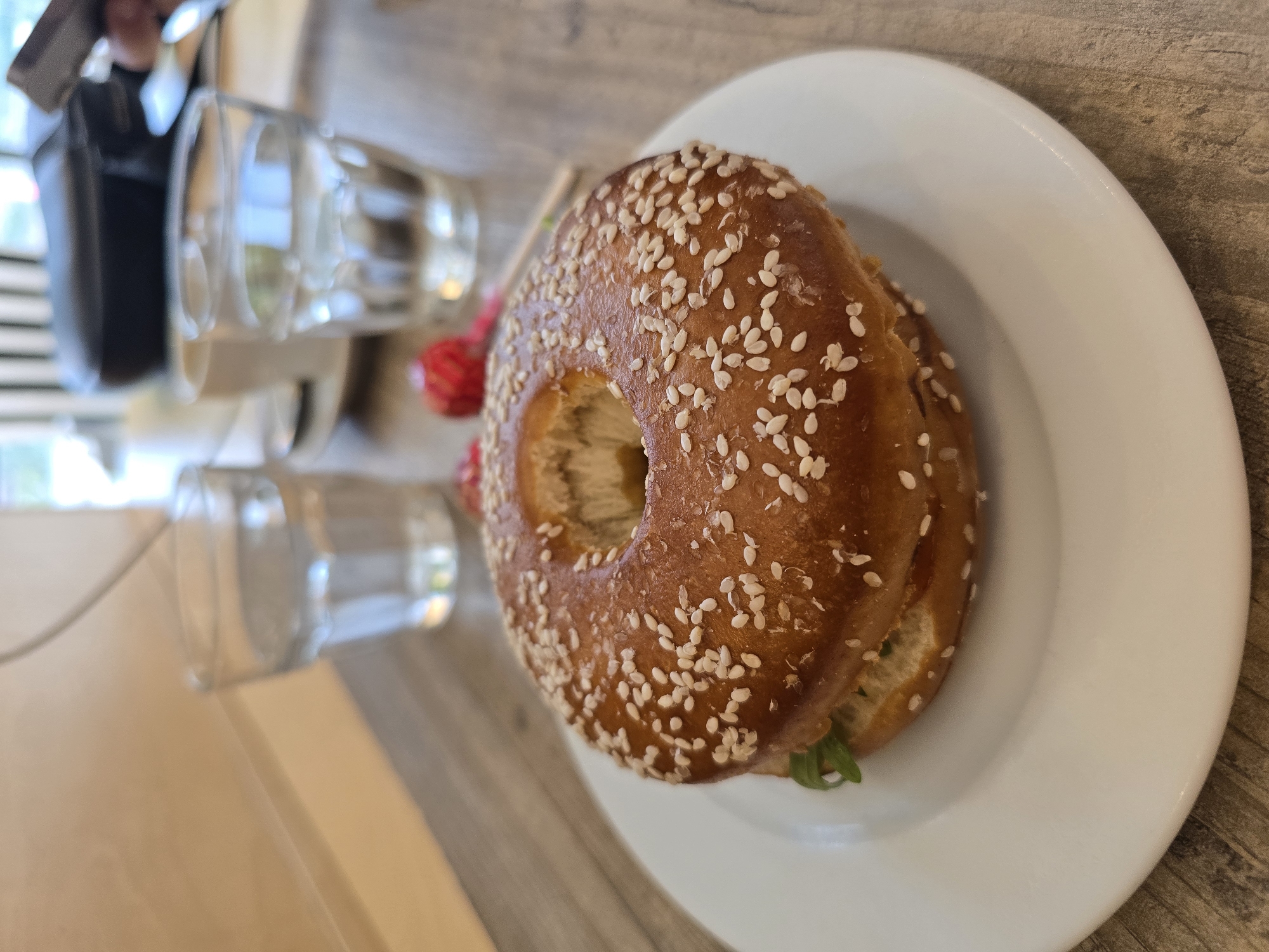 In Baronissi, Italy, on a wooden table, a sesame seed bagel sandwich sits on a white plate. Two glasses of water and a coffee cup are visible in the background. A person's hand holding a phone or tablet is partially visible in the upper right corner. The scene appears to be indoors, possibly a cafe or restaurant, with natural light coming from a window.