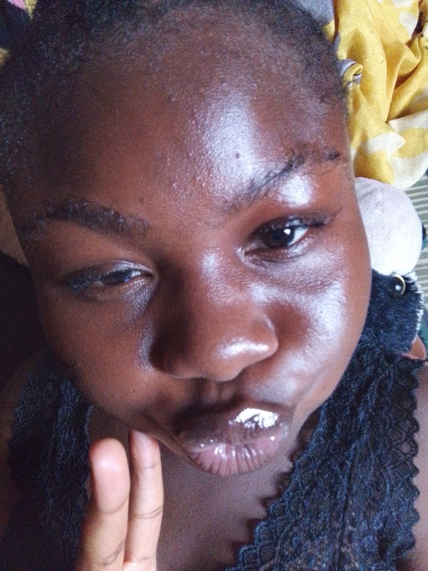 A close-up selfie of a person with dark skin. The person's face fills most of the frame, with the camera positioned above. The person has short, dark hair and dark eyebrows. Their left eye is partially closed. Their lips are shiny. The person's left hand is visible near their chin. A yellow patterned fabric is visible in the upper right corner. A dark-colored, plush object is partially visible behind the person's right ear. The scene is indoors. The image does not provide sufficient details to determine a specific location in Nsukka, Nigeria.