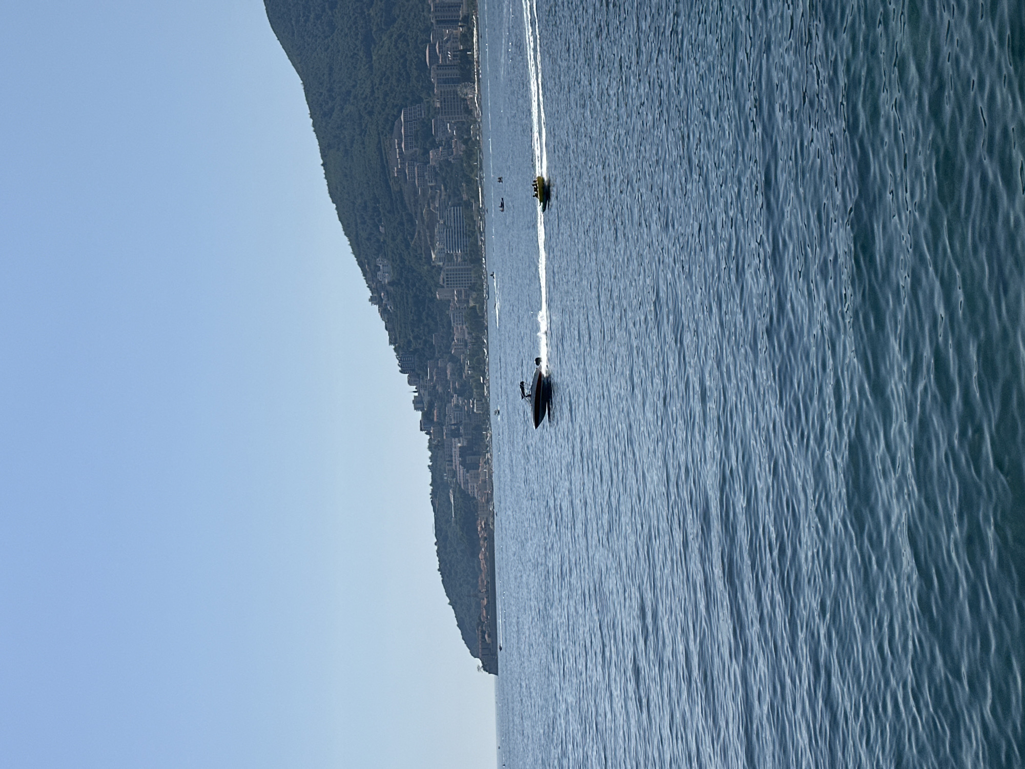 This image captures a panoramic view of the sea in front of the city of Budva, Montenegro. The water is a deep blue, with gentle waves creating ripples across its surface. In the distance, a lush green mountain slopes down towards the coastline, dotted with buildings and structures that form the city. The sky above is a clear, bright blue, typical of a sunny day.
In the foreground of the image, a sleek, dark-colored speedboat is cutting through the water, leaving a white wake behind it. To the right of the speedboat, a yellow inflatable tube is being towed, carrying a few people. They are all enjoying a recreational activity on the water. Several other smaller boats are visible further out in the sea, suggesting a lively atmosphere and the presence of other water activities. The coastline of Budva is characterized by a mix of modern buildings and older structures, with some red-tiled roofs visible amongst the urban landscape. The overall scene depicts a picturesque coastal environment with people engaging in water sports under a clear sky.