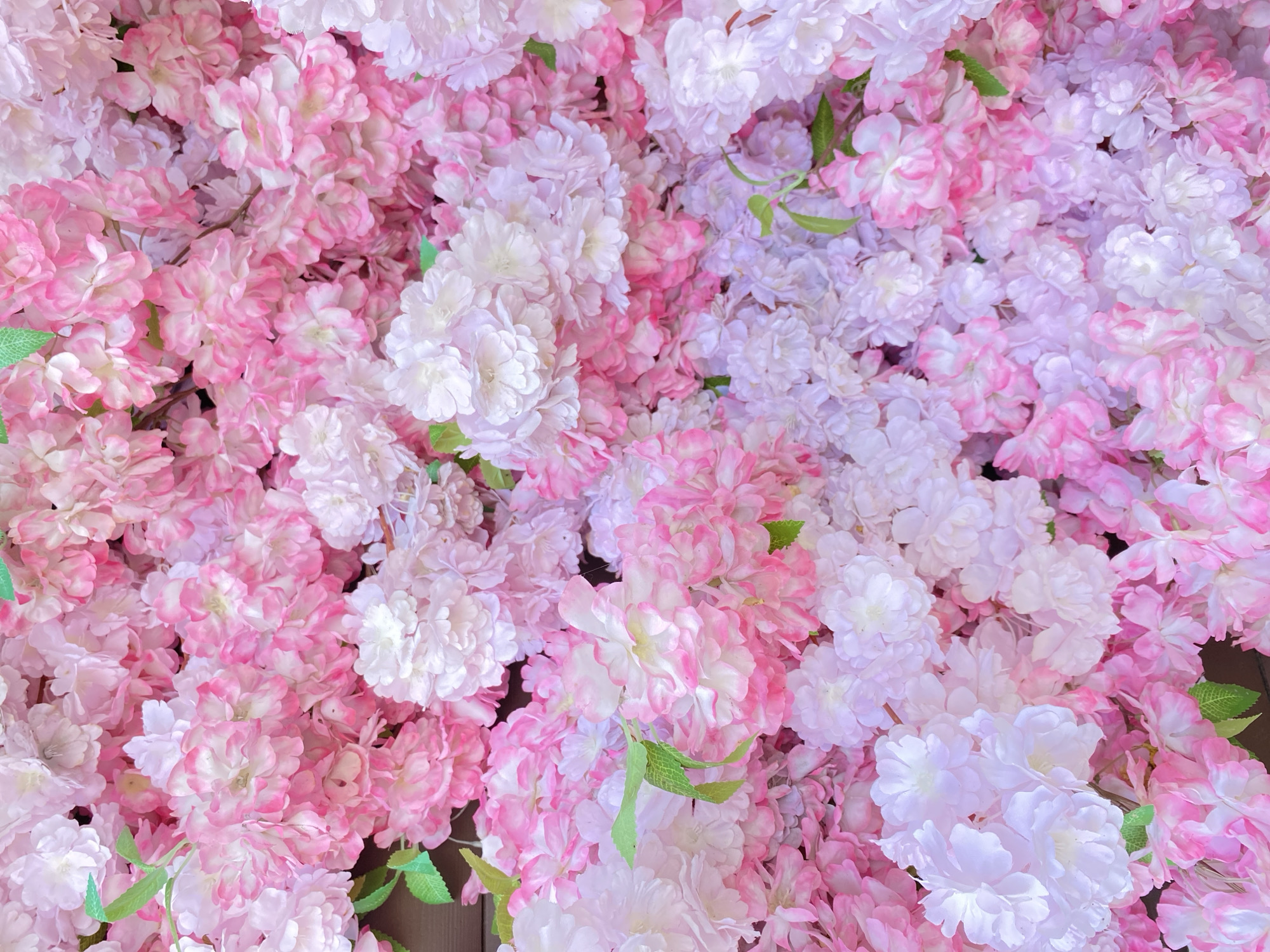 The image displays a dense, full-frame composition of numerous artificial flowers, predominantly in shades of pink, white, and light purple. The flowers appear to be small to medium-sized blooms, each with multiple delicate petals, creating a rich, textured surface.
The dominant color is various hues of pink, ranging from soft pastel pinks to more vibrant fuchsias, often with white centers or edges, giving them a two-toned appearance. Interspersed among these are pure white flowers and others with a light lavender or pale violet tint, adding variation to the floral tapestry. A few small, bright green leaves are visible peeking out from among the blossoms, providing a subtle natural accent.
The flowers are packed closely together, suggesting a floral wall, backdrop, or a large decorative arrangement. There is no discernible activity taking place, and no people or other objects are visible. The lighting is bright and even, highlighting the soft textures and colors of the artificial blooms. The overall impression is one of beauty, softness, and decorative elegance, commonly associated with celebrations, events, or interior decor. No visible text is present in the image.