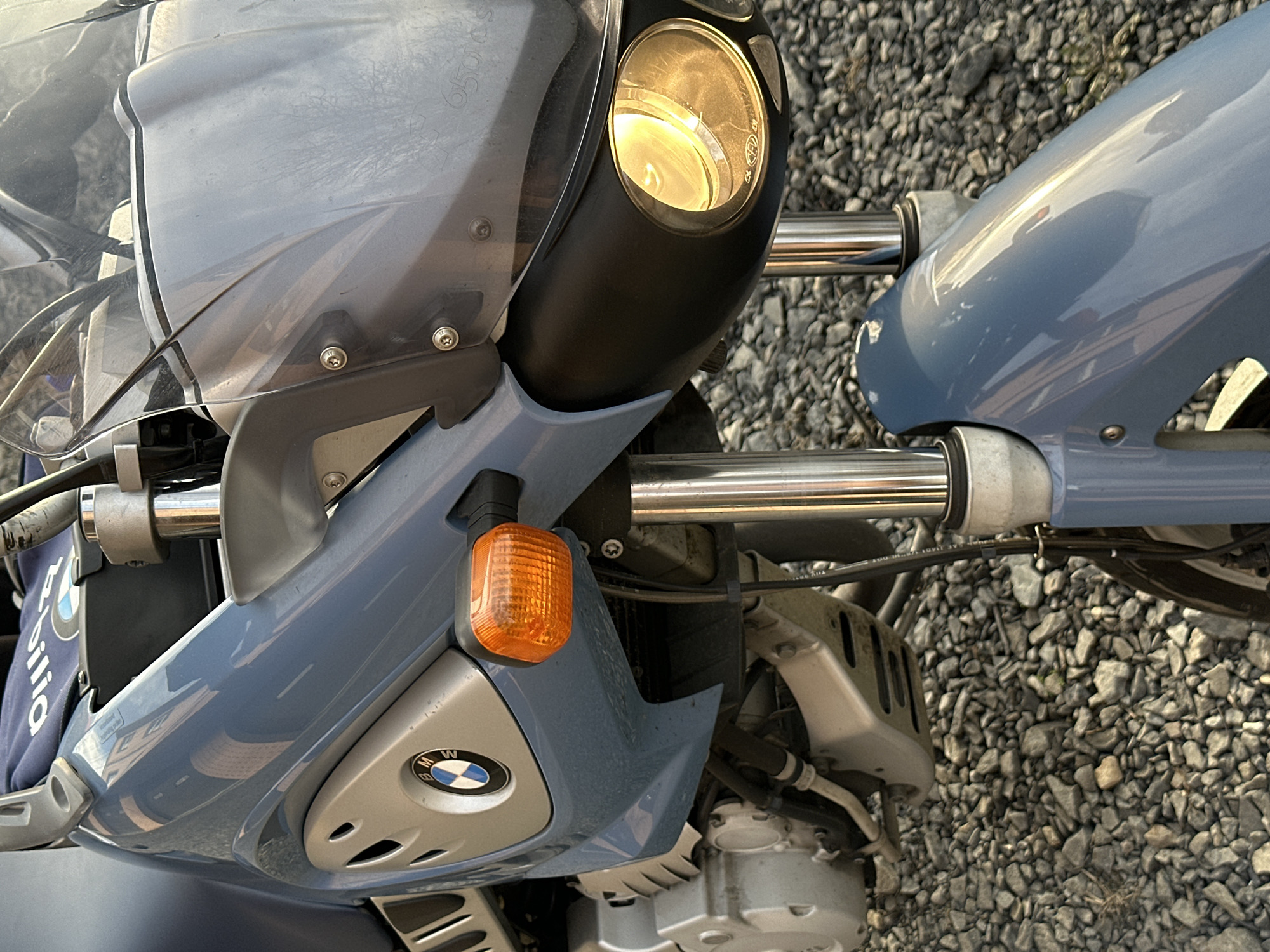 A close-up shot shows the front portion of a light blue BMW motorcycle. The headlight is on, casting a warm glow. An orange turn signal is visible beneath the headlight. The BMW logo is prominently displayed on the front fairing. The motorcycle is parked on a surface covered with small gray rocks. There is no indication of the location being Rostov-on-Don, Russia, or any people, subjects, actions, or interactions in the image.