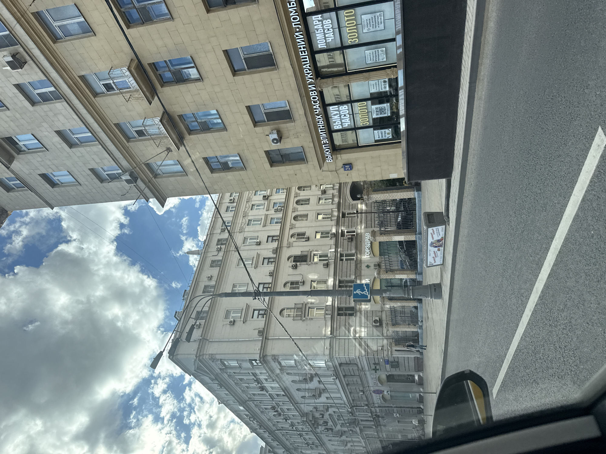 The image captures a street view in Moscow, Russia, likely on a cloudy day. Two multi-story buildings dominate the scene, one a lighter, ornate structure on the left, and the other a yellowish building on the right with multiple windows and balconies. A streetlamp stands prominently near the corner of the buildings. The street in the foreground has asphalt paving with a white line marking.
On the yellowish building on the right, there are several signs. One prominent sign in Russian reads "ВЫКУП ЭЛИТНЫХ ЧАСОВИ УКРАШЕНИЙ - ЛОМБАРД ЧАСОВ" which translates to "PURCHASE OF ELITE WATCHES AND JEWELRY - WATCH PAWNSHOP". Below this, in large lettering, are the words "ЗОЛОТО" (Gold).
On the building to the left, a sign with a blue pedestrian symbol indicating an upward movement is visible, along with a sign that reads "ГОЗДРАВ" (likely a pharmacy chain). A decorative fence runs along the sidewalk in front of this building.
A few individuals are visible on the sidewalk, one walking and another standing near the building. The sky is a mix of blue with significant cloud cover. The perspective suggests the photo was taken from a vehicle.