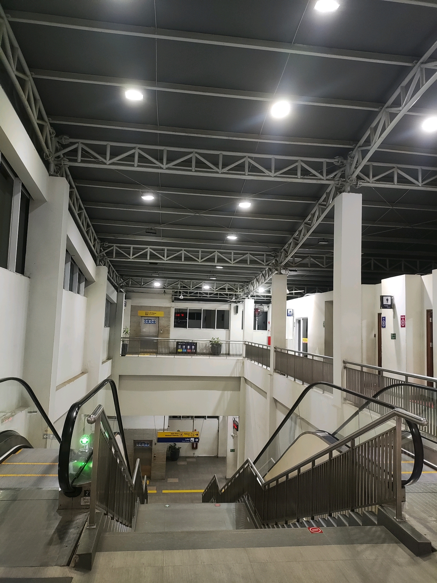 The image shows an interior view of a modern transportation hub, likely a train station or bus terminal, in Telagamurni, Indonesia. The scene is dominated by clean, white walls and a high, industrial-style ceiling with exposed metal trusses and fluorescent lighting.
In the foreground, a wide concrete staircase descends from the viewer's perspective, flanked by metal railings. To the left of the stairs, an escalator moves downwards, its black handrail and metallic steps clearly visible. The escalator has a green illuminated indicator at its base.
Further into the scene, a mezzanine level is visible, also with metal railings. On this level, there are signs, including a yellow sign with "Lift Keluar" (Exit Elevator) and "Jalan Raya A" (Main Road A) indicating directions. There are doorways and what appear to be public restrooms, identifiable by pictograms of a male figure and a female figure above their respective doors. A red fire extinguisher is mounted on the wall near one of the restrooms.
The environment is well-lit and appears to be clean and orderly. No people are visible in the immediate vicinity of the stairs or escalators. The overall impression is that of a functional and contemporary public space.