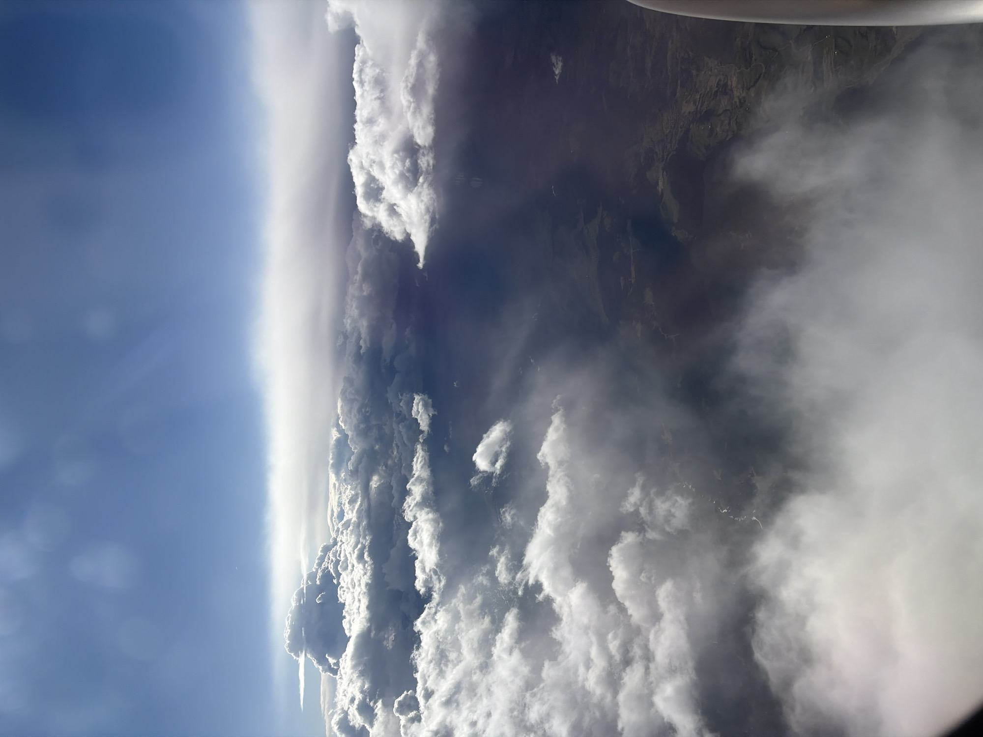 An aerial view, likely taken from an aircraft, shows a stark division between a clear blue sky and a dense cloud formation that obscures much of the landscape below. The clouds, ranging from light white to dark grey, create a turbulent horizon. Some land is visible through gaps in the clouds, hinting at a mountainous terrain, though its specific features are unclear due to the cloud cover. No people, actions, or interactions are visible. The location, Babaići, Montenegro, is not visually verifiable from the image itself but is provided as additional information.