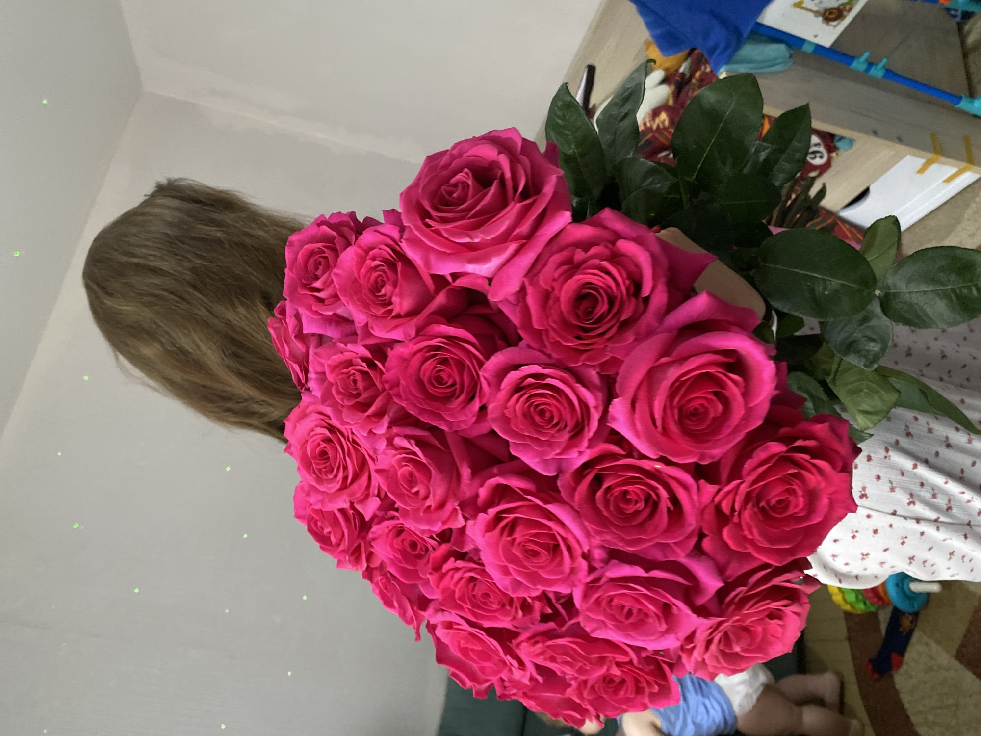 A person with light brown hair holds a large bouquet of bright pink roses with green foliage. The backdrop includes a light-colored wall and a room interior with a child in blue clothing lying on the floor. The location is likely indoors, potentially a residence in Votkinsk, Russia. The scene features an adult holding the bouquet of roses.