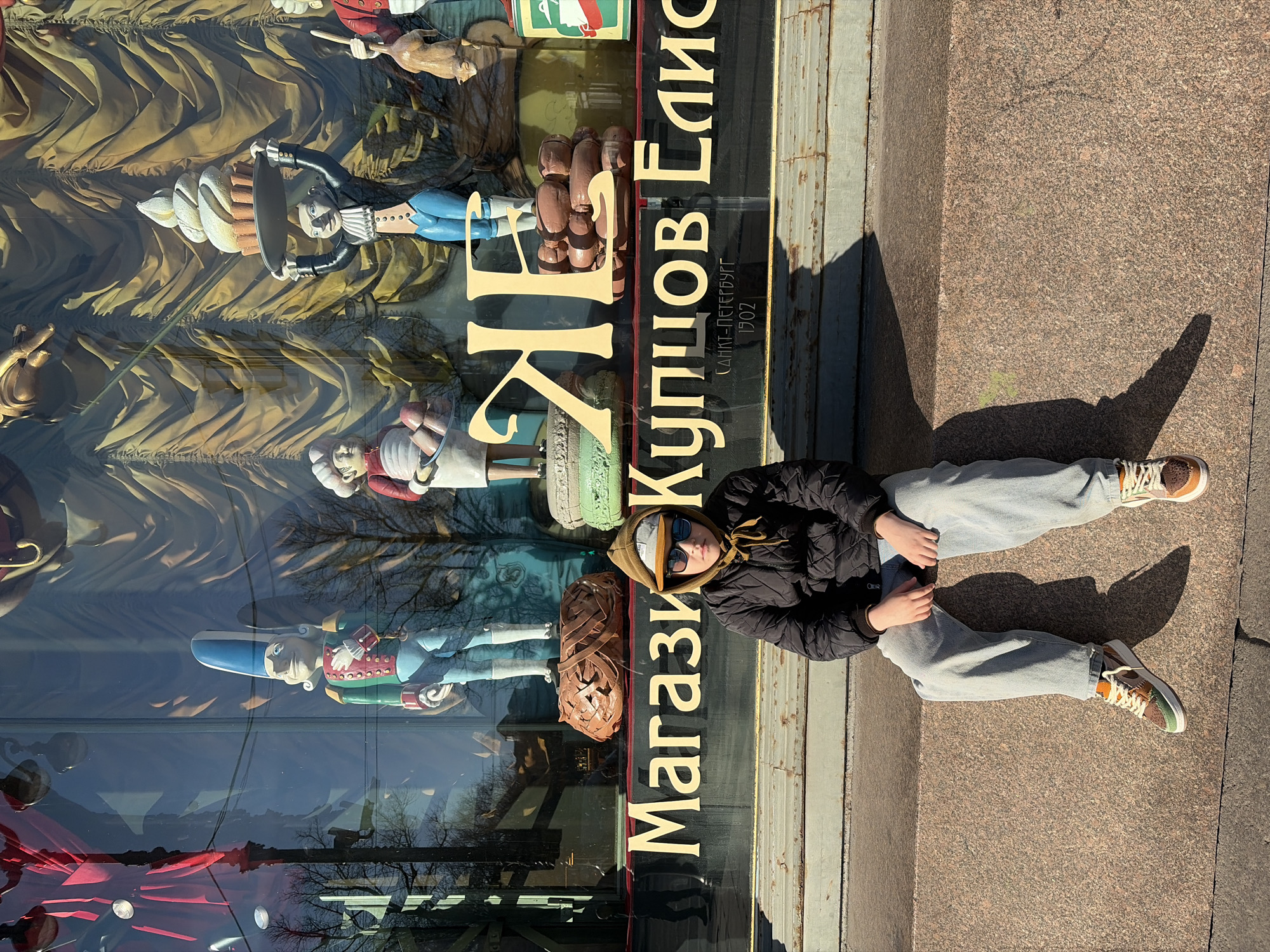 A person is sitting on a concrete surface next to a shop window in Saint Petersburg, Russia. The shop window is for "Магазин Купцов Елисеевых" (Eliseev Emporium), as indicated by the gold lettering on the glass. The letters "САНКТ-ПЕТЕРБУРГ" and "1902" are also visible. The window displays a variety of decorative items including what appear to be figurines of people and confectionery. The person sitting is wearing a dark jacket, grey sweatpants, and sneakers. They are also wearing sunglasses and a hood. The person is facing the shop window and is in a relaxed position. The ground is made of a rough, granite-like material and a long shadow stretches across it from the seated person.