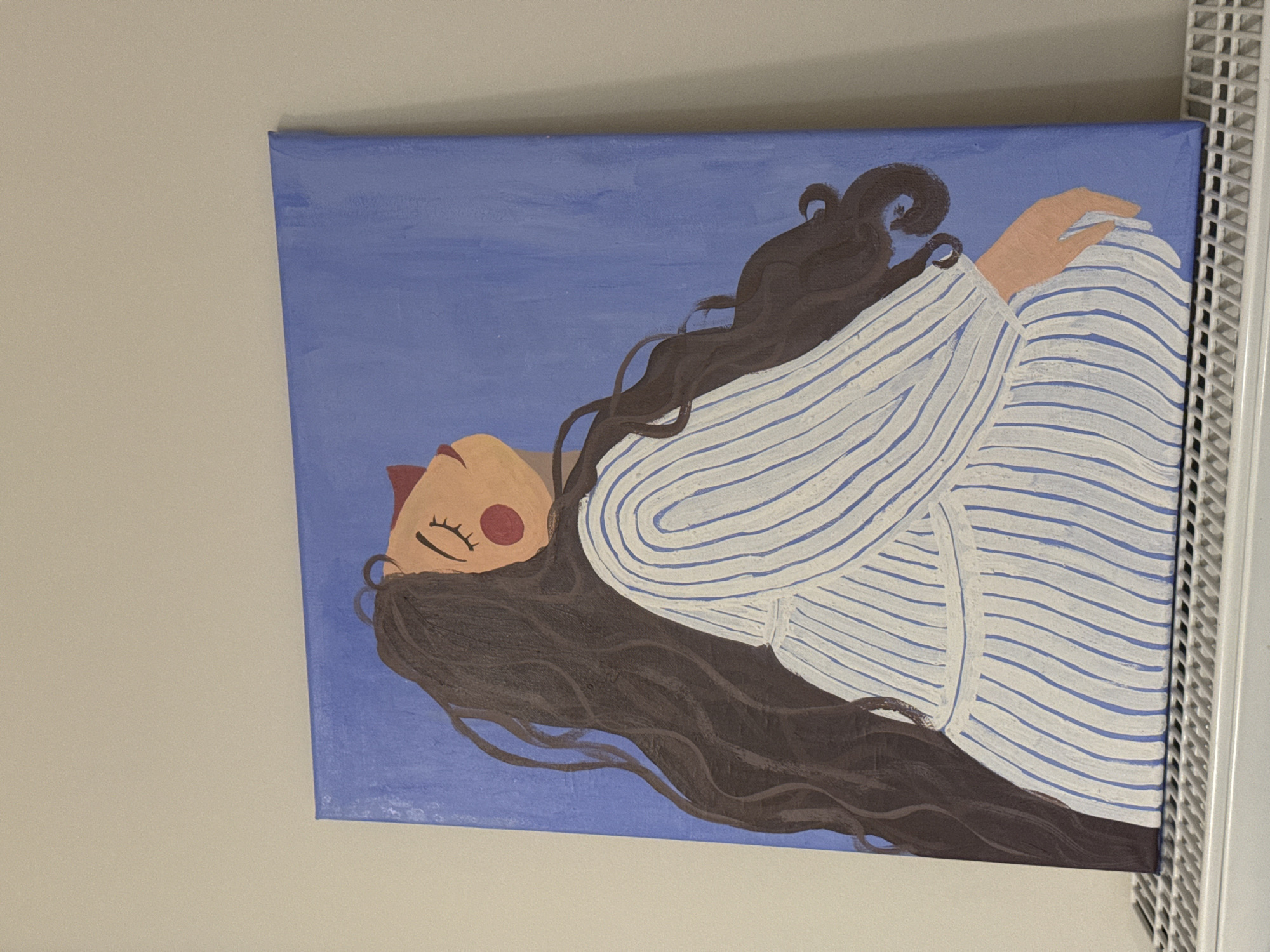 A painting of a woman is shown, tilted on its side against a light beige wall. The woman is depicted with her eyes closed, suggesting rest or sleep, and a serene expression with slightly parted reddish-brown lips and rosy red cheeks. Her long, dark, wavy hair flows around her head. She has a light peach skin tone.
The woman is wearing a white top with irregular horizontal blue stripes, and one hand is visible, resting near her head. The background of the painting is a solid periwinkle blue. The art style appears hand-painted, with a somewhat stylized and folk-art aesthetic, characterized by soft lines and distinct color blocks.
To the right of the canvas, a white slatted object, possibly a shelf or vent, is partially visible. The overall scene suggests an indoor setting, with the painting displayed on a wall. There is no visible text in the image.