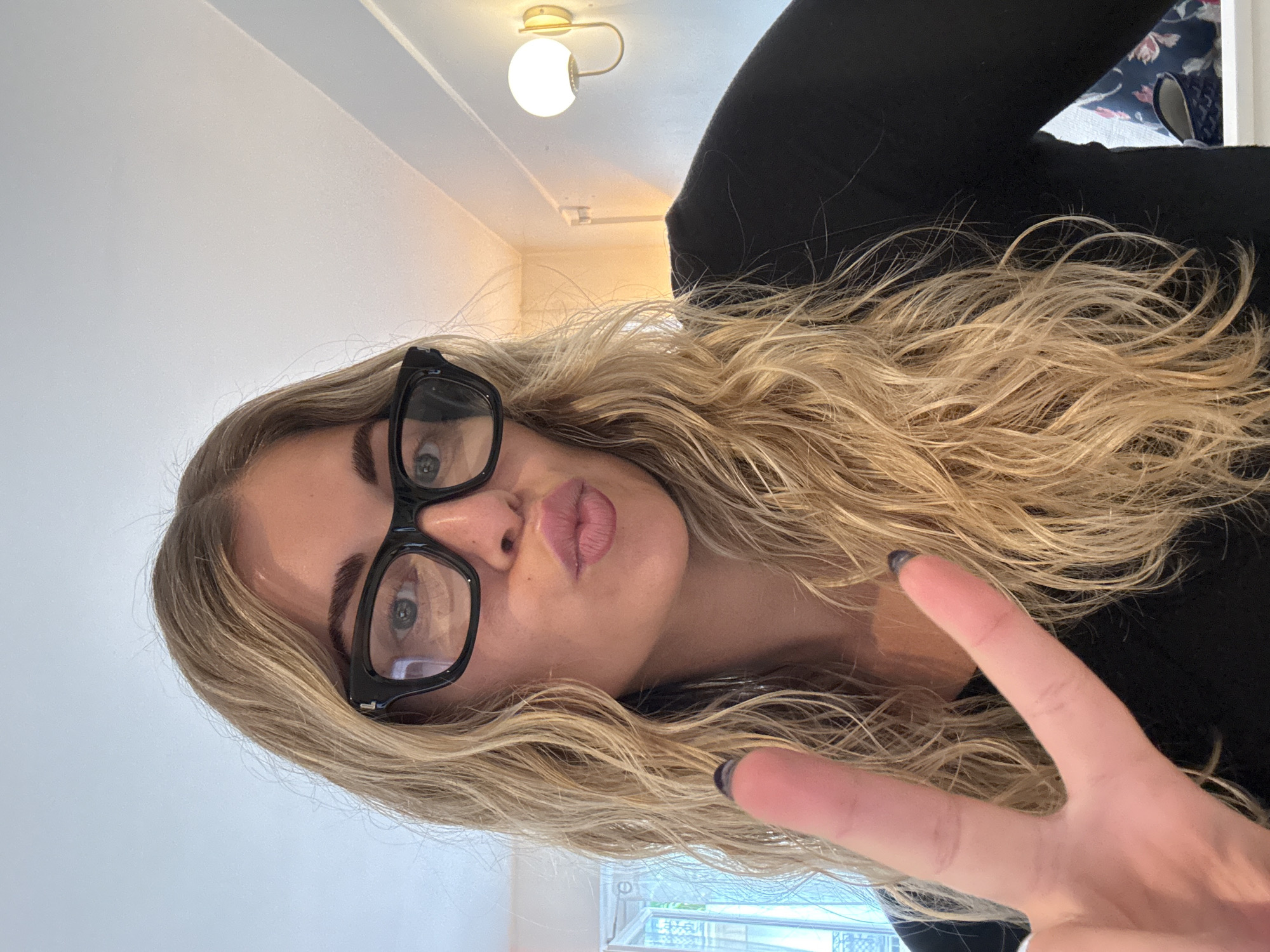 A woman with long, wavy blonde hair, wearing black-framed glasses, is making a "peace" sign with her hand and pursing her lips towards the camera. The background includes a light fixture, a white wall, and a glimpse of what appears to be a window with a view that may be consistent with a Parisian balcony or window box. Without more information, I cannot provide a specific location.