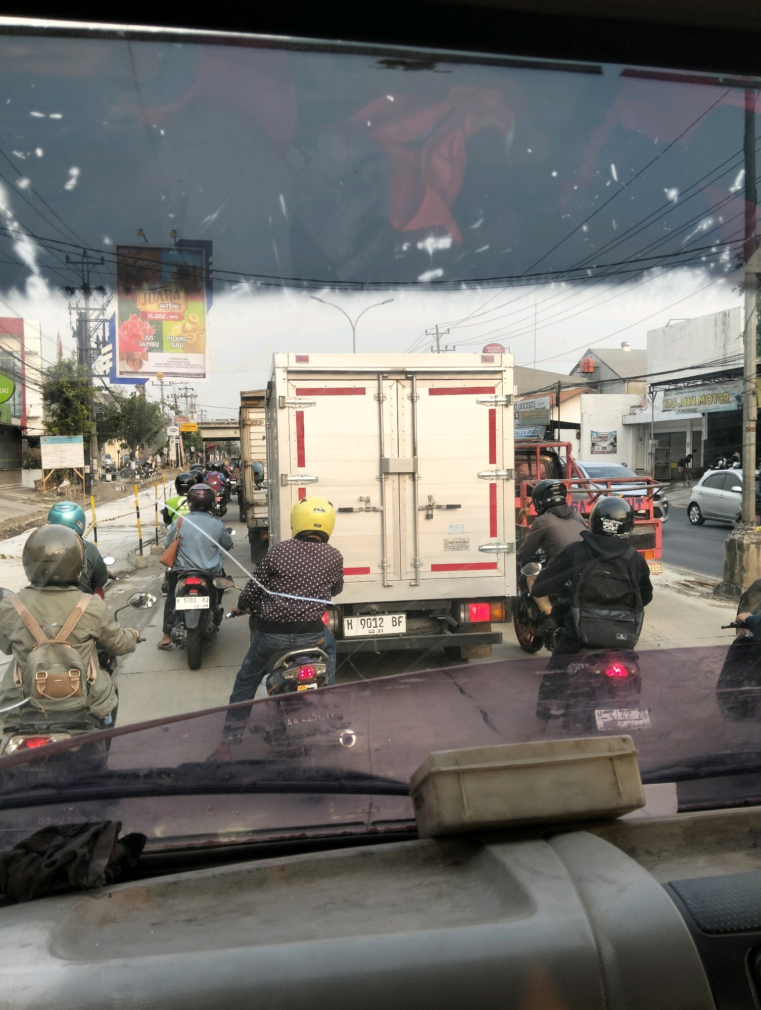 In Semarang, Indonesia, a traffic jam is captured on a cloudy day, viewed from inside a vehicle. The foreground shows the vehicle's dashboard and windshield, with a discarded cloth lying on the dashboard. Beyond the windshield, a line of motorcycles and a truck are visible. On one motorcycle, a person wearing a yellow helmet and a polka-dot shirt is seen from behind. Another person in a green helmet and a brown backpack is riding a motorcycle in front of them. A white truck with red accents occupies a significant portion of the mid-ground, its rear doors closed. Several other motorcycles are interspersed in the traffic. In the background, buildings line the street, and power lines crisscross overhead. A billboard advertising "JUARA INTENS" for juice and bananas is also visible. The overall scene depicts a typical urban traffic situation with various modes of transportation.