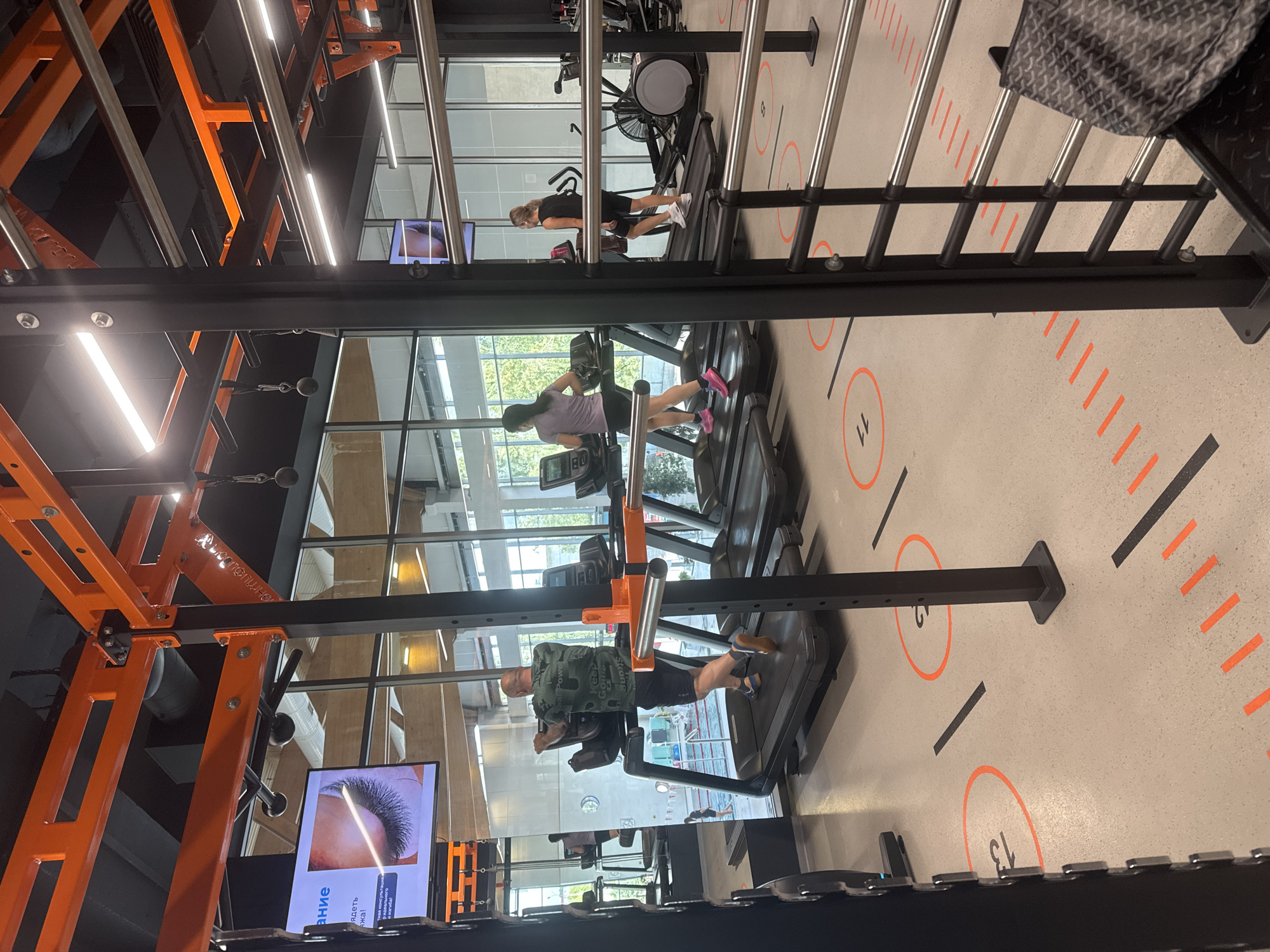 The image depicts the interior of a modern gym, likely located in Sovkhoza imeni Lenina, Russia, as suggested by the contextual information. The scene is dominated by exercise equipment, particularly treadmills. In the foreground, a man with a shaved head is walking on a treadmill, wearing a green t-shirt with a repeating pattern of white text and dark shorts. To his right, further back, two women are also on treadmills. One woman, with dark hair pulled back, wears a light purple t-shirt and black shorts. The other woman, with blonde hair, wears a black sleeveless top and black shorts.
The gym features an industrial aesthetic with exposed orange metal beams and black structural elements. Large windows line the back wall, offering a view of an outdoor swimming pool area and some greenery, indicating the gym might be part of a larger recreational complex. The floor is a light grey speckled surface with painted orange circles and black lines, likely designating workout zones or spacing markers. A large screen displaying an image of an eye with long lashes is visible on the left side of the image, mounted high on a wall. Other gym equipment, including what appears to be an air bike and part of a weight rack, can be seen in the background. The lighting is a mix of overhead fluorescent lights and natural light from the windows.