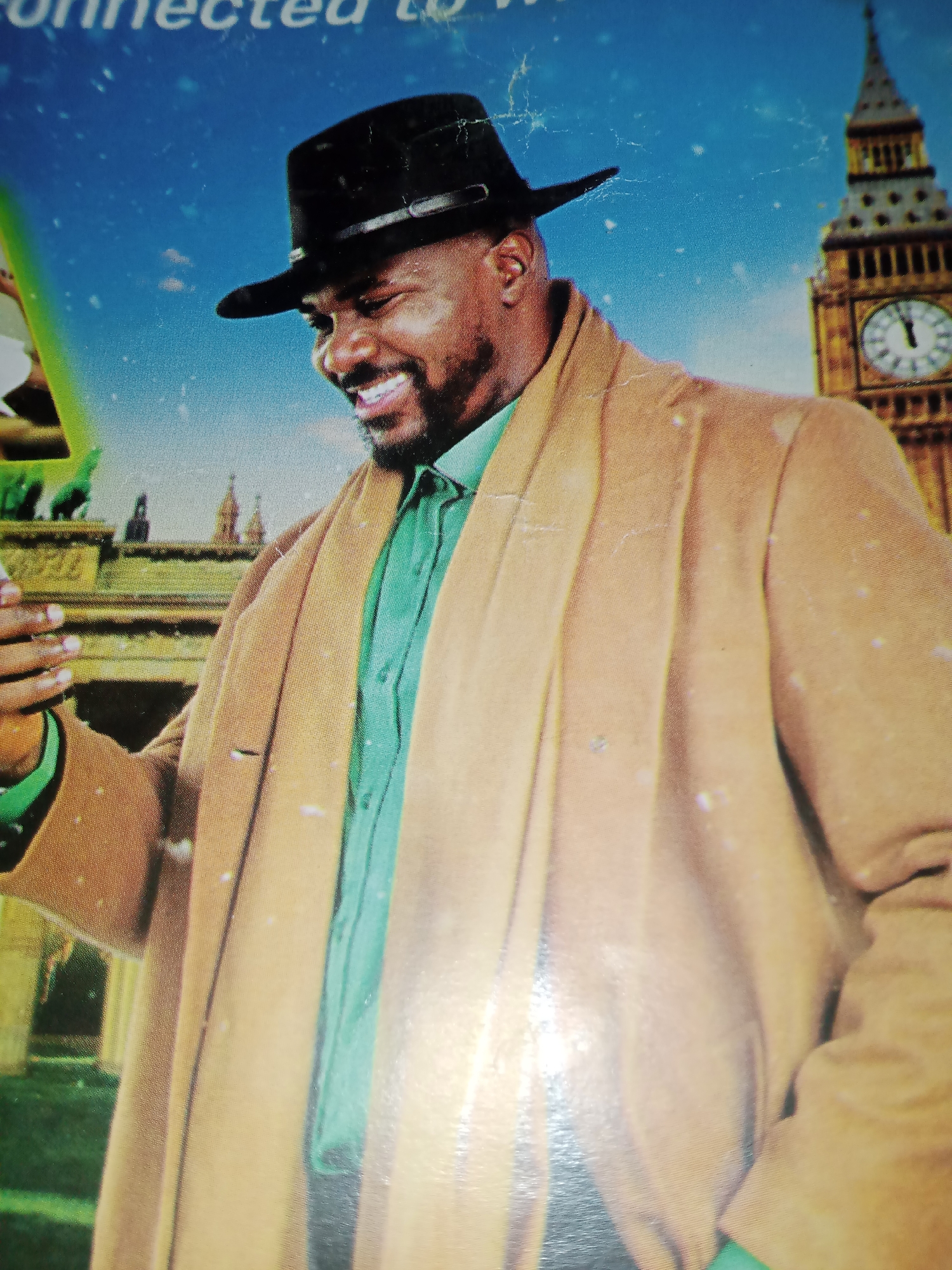 The image is a close-up photograph of a man wearing a black fedora, a green collared shirt, and a tan overcoat. He is smiling and looking down to his left, with his right hand raised near his face. In the background, out of focus, are elements of a city scene, including a tall clock tower that resembles Big Ben, other buildings, and a snowy sky. The man's attire and the background suggest a European setting, likely London, rather than Jalingo, Nigeria. The image appears to be a still from a movie or advertisement.