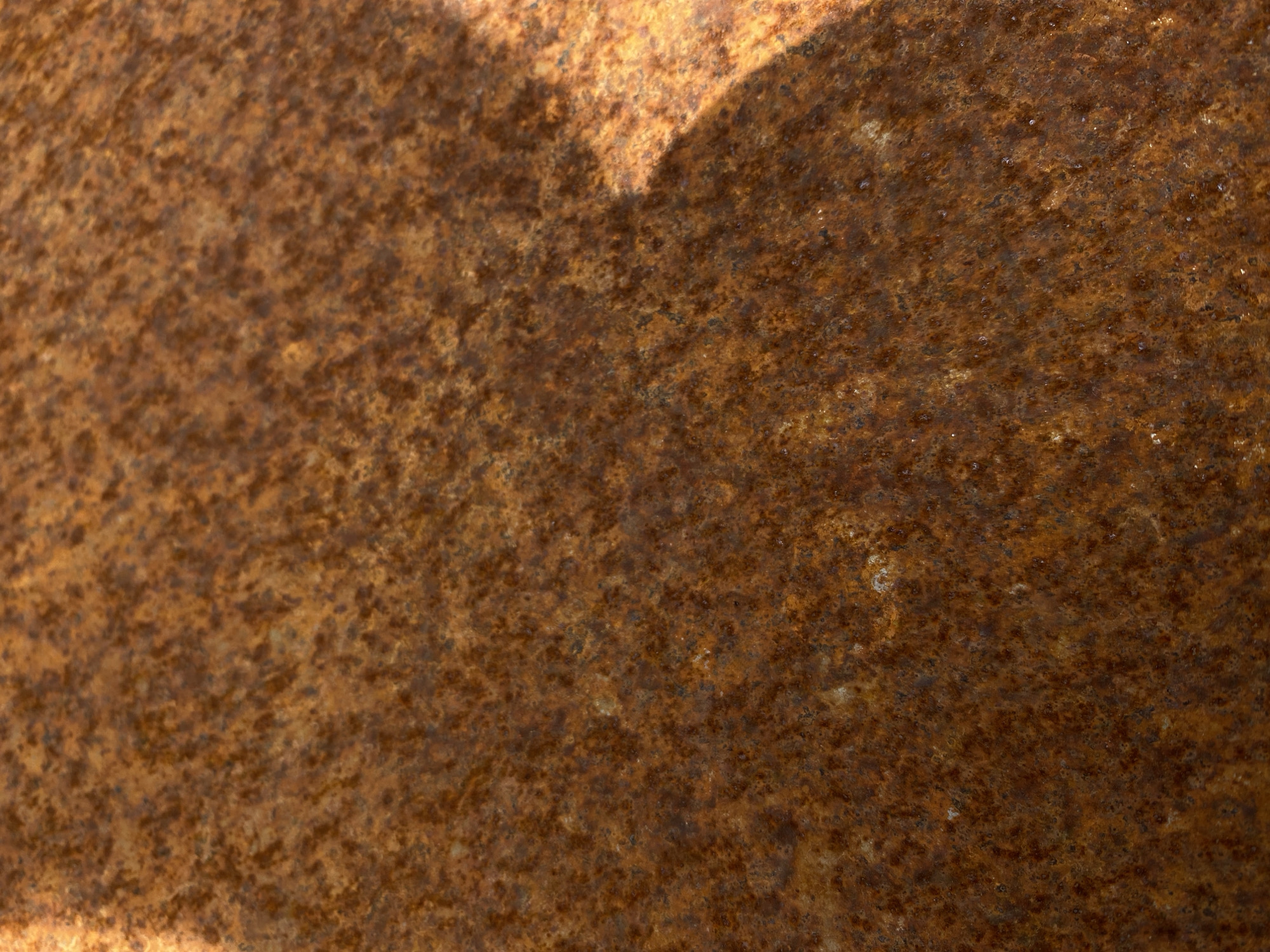 The image shows a close-up, textured view of a rusty metal surface. The rust is predominantly in shades of brown and orange, with some darker patches and lighter, almost yellowish areas. The texture appears granular and uneven, with small pits and raised areas. There is a shadow cast on the right side of the image, suggesting an external light source and an object casting the shadow, although the object itself is not visible. The lighting creates a slight sheen on the surface. There are no people, animals, or specific objects identifiable beyond the rusty metal itself. The context provided does not contain information about the location in the city or any specific environment other than the material itself.
