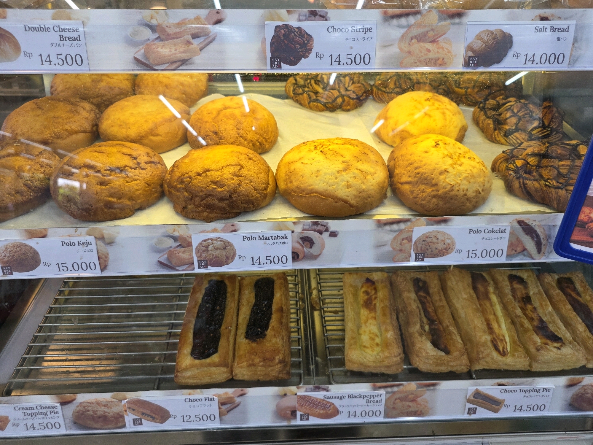 The image shows a display of baked goods in a store, likely a bakery or a supermarket, in Tanjung Pinang, Indonesia. The display is organized into shelves filled with various types of bread and pastries. Each item has a label indicating its name and price in Indonesian Rupiah (Rp).
On the top shelf, there are several round, golden-brown breads labeled "Double Cheese Bread" (Rp 14.500), "Choco Stripe" (Rp 14.500), and "Salt Bread" (Rp 14.000). Behind these, some pastries with chocolate swirls are visible, possibly the "Choco Stripe" bread.
The second shelf displays more round, golden-brown breads and pastries. Labels visible are "Polo Keju" (Rp 15.000) and "Polo Martabak" (Rp 14.500). Some of these appear to be filled.
The third shelf showcases rectangular pastries, some with dark filling and others with a lighter, possibly cheese filling. Labels include "Cream Cheese Topping Pie" (Rp 14.500), "Choco Flat" (Rp 12.500), and "Sausage Blackpepper Bread" (Rp 14.000). Another label for "Choco Topping Pie" (Rp 14.500) is partially visible on the right.
The environment appears to be an indoor retail space with bright lighting, highlighting the products. The shelves are made of metal grates, and the display case is made of glass. No people are visible in the frame. The overall impression is one of abundance and variety of baked goods.