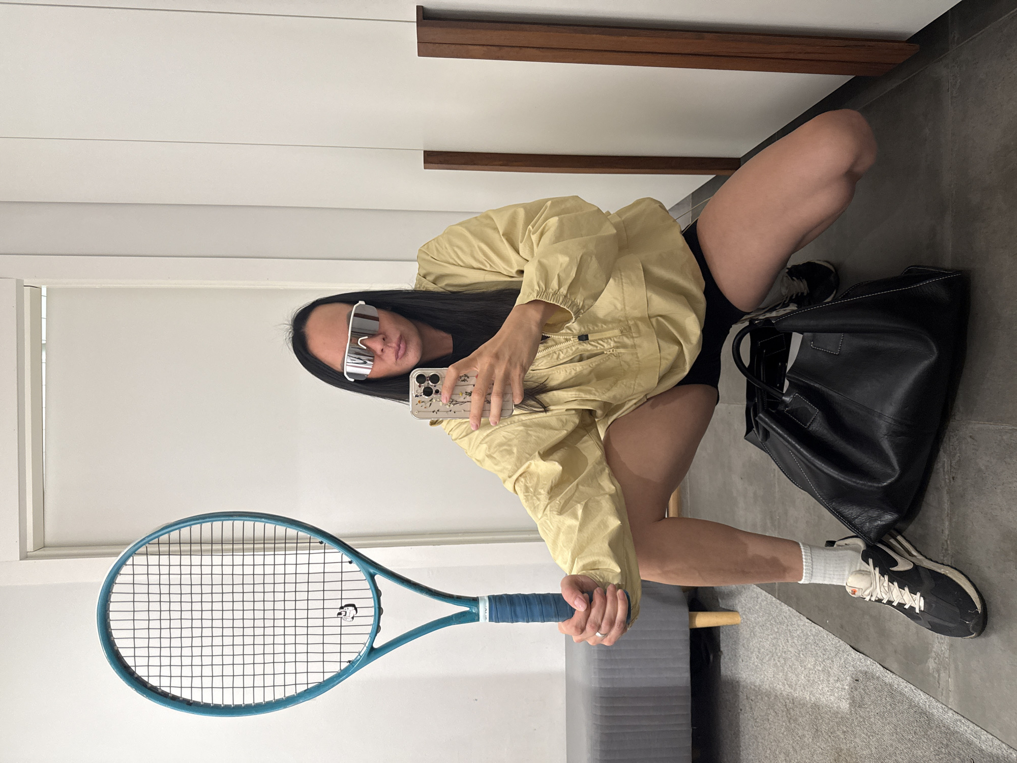 A person is in a squatting position, holding a tennis racket. They are wearing a light yellow jacket, black shorts, white socks, and black and white sneakers. They are also wearing white wrap-around sunglasses and have long, dark hair. The person is taking a selfie with a smartphone. In the foreground, a large black bag sits on the floor. The background consists of white walls and a doorway. The person's left hand is wearing a ring. The image appears to be taken indoors, possibly in a locker room or changing area, but its exact location, including in the city of Chaikovsky, Russia, cannot be determined from the visual information provided.