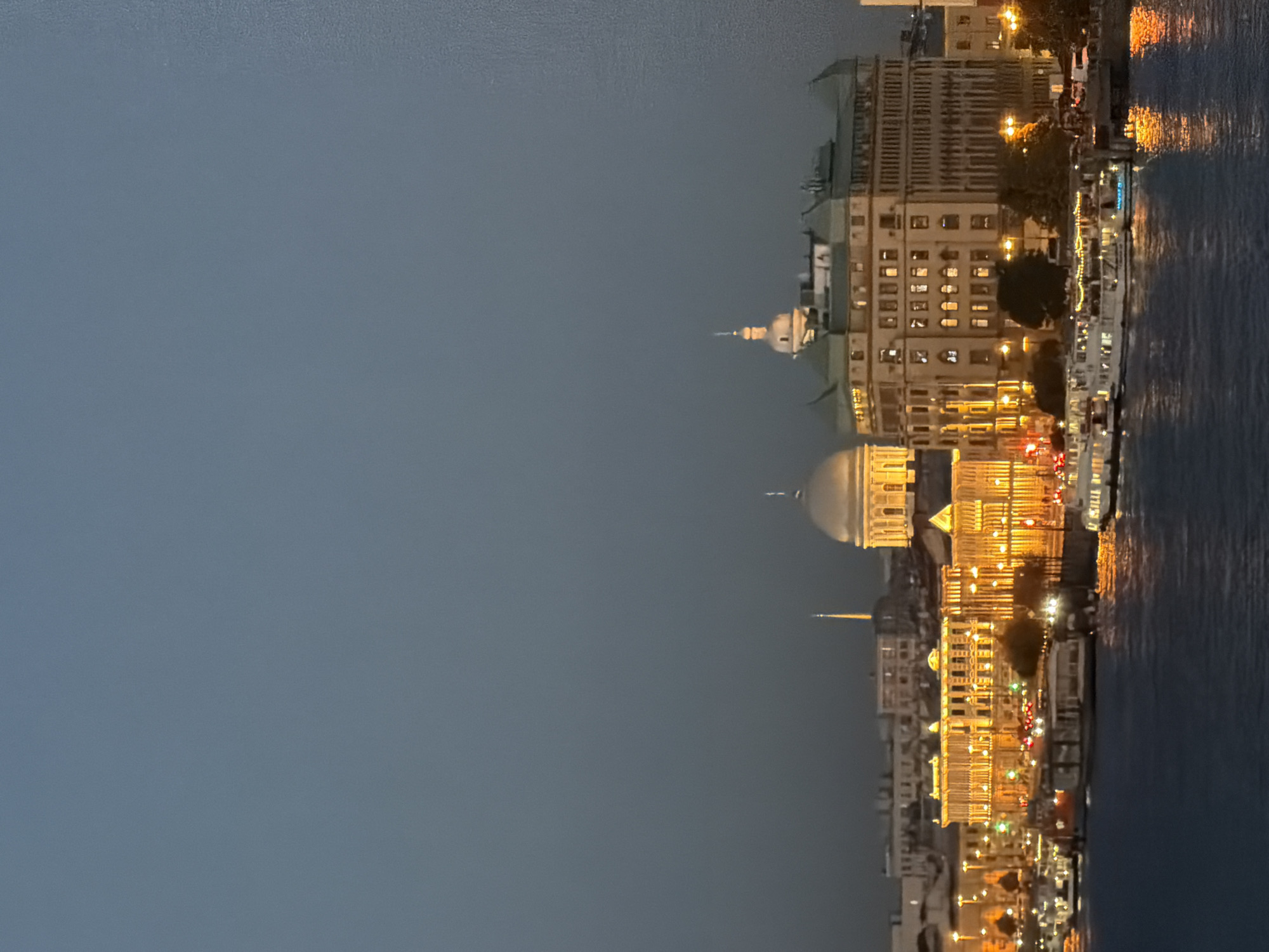 The image captures a nighttime view of the Saint Petersburg cityscape along the Neva River. Multiple illuminated buildings with classical architecture line the riverbank, their warm yellow lights reflecting on the dark water. Prominent structures include a large building with a dome and a smaller, pointed spire. Several boats and ferries, also lit up, are docked or moving on the river. The sky above is a deep, muted blue, suggesting twilight or a heavily overcast evening. The overall scene evokes a serene and majestic ambiance of the city at night.