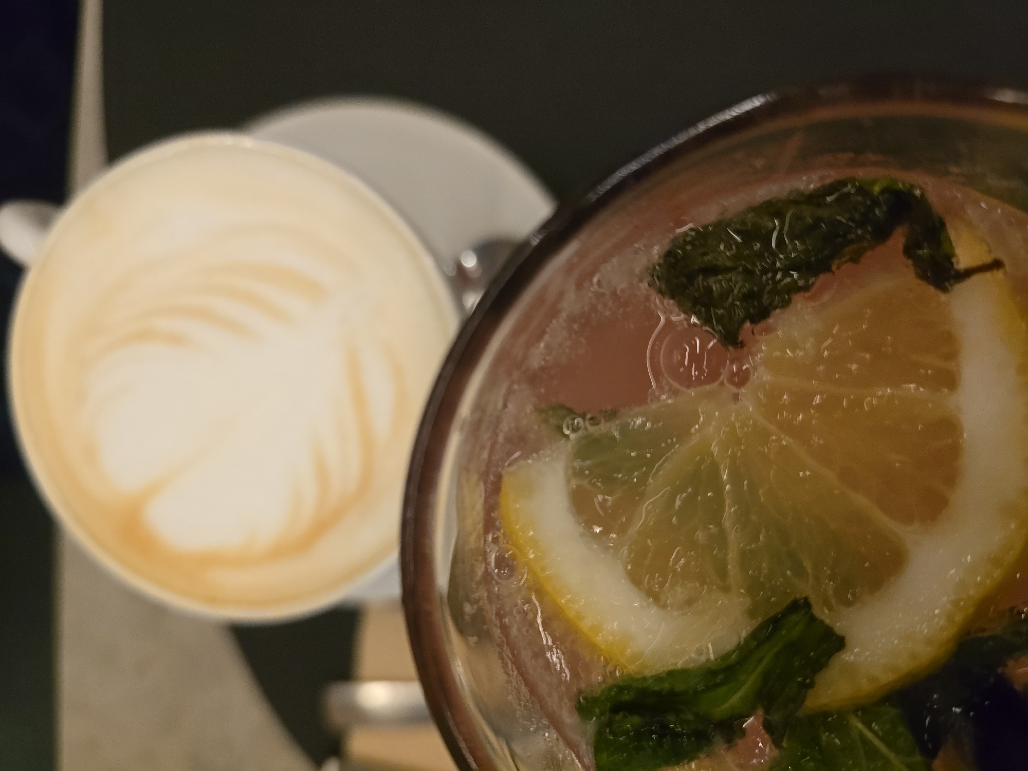This top-down, close-up image captures two distinct beverages set on a surface in a dimly lit indoor environment, likely a cafe or restaurant in Varna, Bulgaria.
On the left, slightly out of focus, is a white ceramic cup with a matching saucer. The cup contains a light-colored coffee beverage, most probably a latte or cappuccino, distinguished by its intricate, creamy latte art featuring a swirling pattern on its surface.
Taking prominence on the right side of the frame, and in sharper focus, is a clear glass filled with a vibrant, pinkish liquid. This refreshing drink is garnished generously with several bright yellow lemon or lime slices and fresh green mint leaves. Small bubbles are visible on the interior surface of the glass, suggesting it is a cold or carbonated beverage, such as a flavored lemonade, spritzer, or a fruit-infused cocktail.
The lighting is soft and artificial, creating a cozy ambiance and highlighting the details of the drinks against a dark, blurred background. The scene evokes a quiet, relaxed moment, possibly during an evening outing. No people or visible text are present in the image.