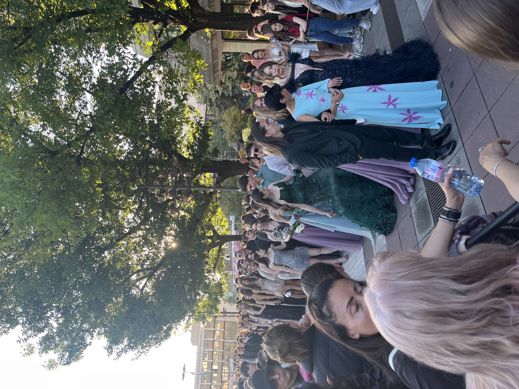 The image shows a large group of people, mostly young women, lining a paved area outside the American University in Blagoevgrad, Bulgaria. Many are dressed in formal gowns. Some individuals are in street clothes or dark suits. Some women have flowers, likely bouquets. A few individuals are holding bottled water. A tall tree casts a shadow over part of the group. A building with Cyrillic script is visible in the background.