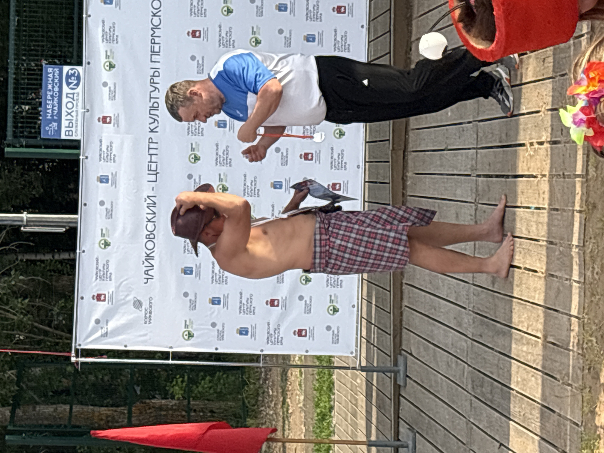 The image shows two men on a wooden outdoor stage in front of a large banner. The banner has Cyrillic text that appears to be promotional or informational, with logos and repeating patterns. On the left side of the banner, there's a sign in Russian that reads "НАБЕРЕЖНАЯ Г.ЧАЙКОВСКИЙ ВЫХОД №3 СЛУЖЕБНЫЙ ПРОЕЗД" which translates to "Chaikovsky Embankment Exit No. 3 Service Drive".
The man on the left is wearing a blue and white t-shirt and black pants. He is bending over, holding a red string with a white object at the end, possibly a game or a prop. The man on the right is wearing a plaid skirt and no shirt. He is wearing a hat and holding a book or folder. His legs are bare.
The overall scene appears to be an outdoor event, possibly a festival or performance, given the stage and the banner. The lighting suggests it is daytime, with sunlight casting shadows on the wooden planks of the stage. The presence of a red flag at the bottom left further suggests an organized event.