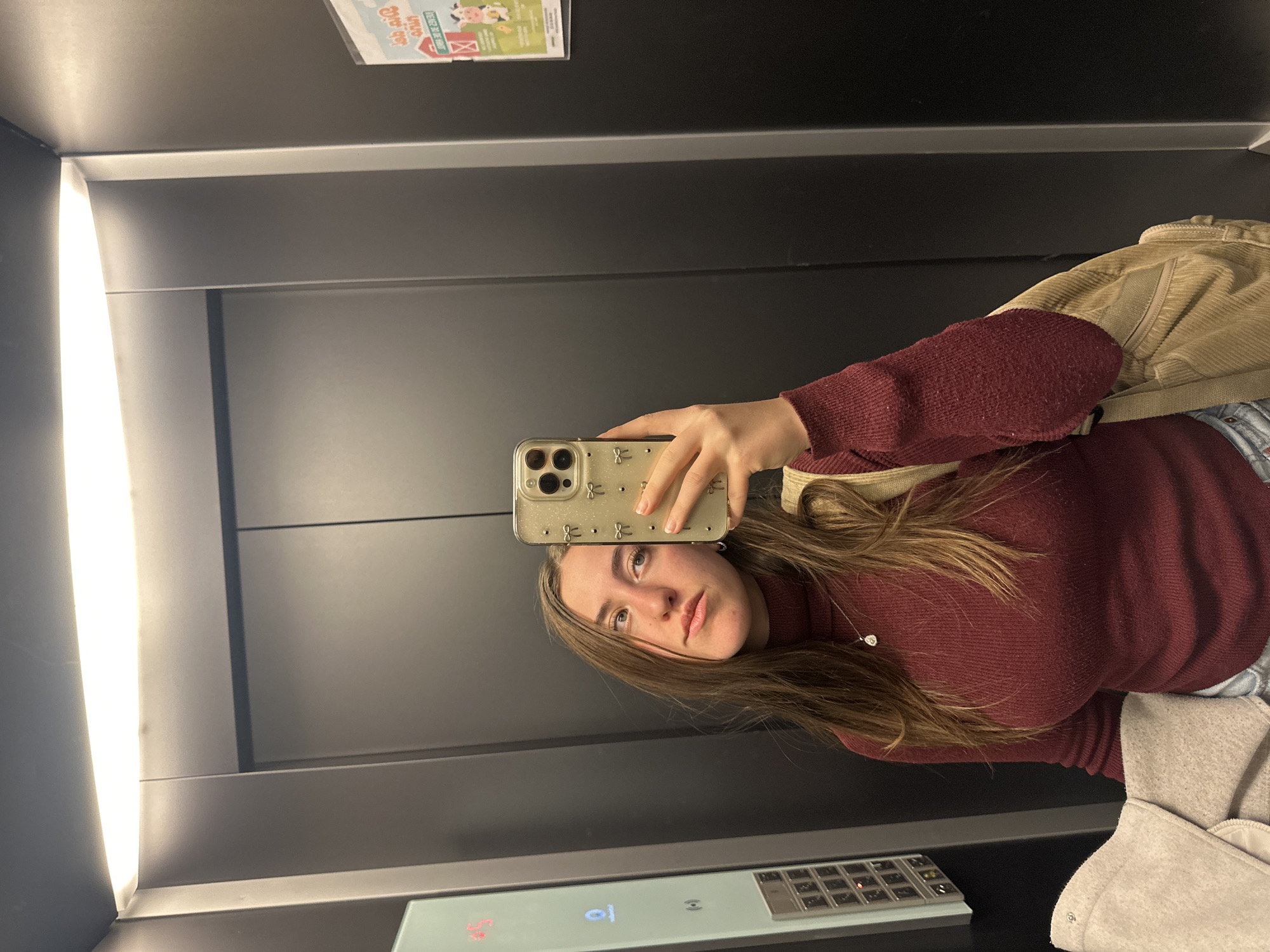 A young woman is depicted taking a selfie in an elevator, holding her smartphone up to capture her reflection. She has long, straight brown hair and wears a maroon turtleneck sweater, light blue jeans, and a delicate silver pendant necklace. A light-colored, possibly corduroy, jacket or bag strap is visible over her right shoulder, and another piece of light beige/grey fabric is seen in the bottom right. Her expression is neutral and slightly contemplative.
The elevator interior features dark, reflective metallic walls, a bright vertical strip of artificial light on the left, and a control panel on the lower right. The control panel clearly shows the number "2" illuminated in light blue, indicating the current floor or destination. The woman's smartphone has a light-colored case adorned with a repeating pattern, possibly bows or small figures, and its multi-lens camera array is prominent.
In the upper right corner, a mirrored reflection of a small poster is visible on the elevator wall. Despite being reversed, legible text includes the Spanish words "GRANJA" (Farm) and "EDUCAR ES JUGAR" (To Educate Is To Play), alongside a graphic depicting a cow and a barn. The scene is brightly lit by the elevator's internal lighting. The image's contextual location is Huixquilucan de Degollado, Mexico.