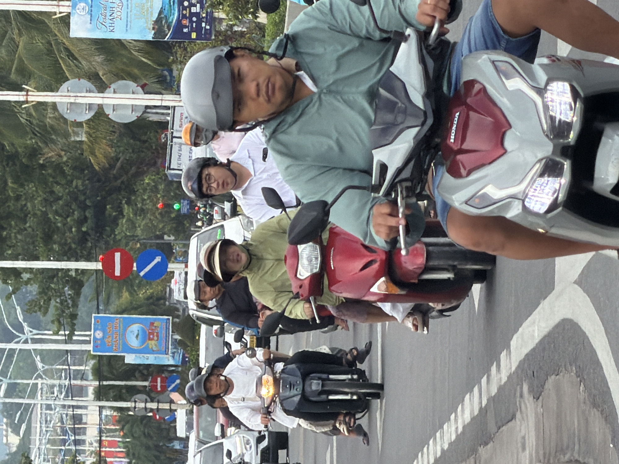 In Nha Trang, Vietnam, a group of people are riding motorcycles down a city street. The image captures a traffic scene with several motorcycles in motion. In the foreground, a person wearing a light blue jacket and denim shorts rides a silver and red Honda scooter. They are looking forward with a neutral expression. Behind them, a person in a green jacket is on a red motorcycle, their face obscured by a hat. Further back, a person in a white shirt and dark pants rides a black motorcycle, also wearing a helmet. The street is paved and has lane markings. Traffic lights are visible in the background, indicating an intersection. Buildings and palm trees line the street, suggesting an urban environment. Several advertising banners and street signs are also present, including one with Vietnamese text and a picture of a boat. The sky is overcast.