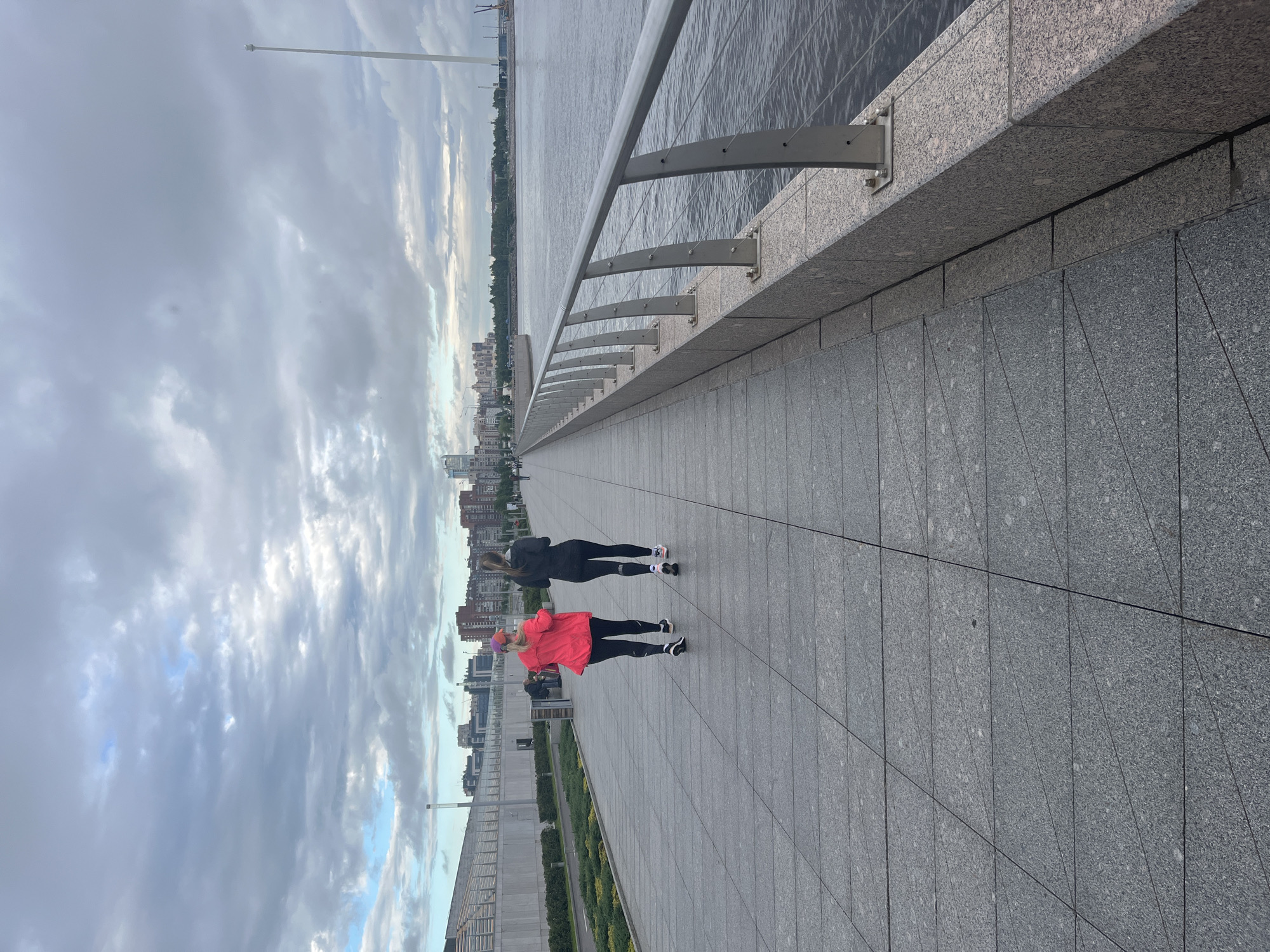 The image depicts two women jogging along a wide paved walkway beside a body of water. The walkway is bordered by a modern railing and a wall made of large granite tiles. In the background, a city skyline with numerous buildings is visible, and the sky is overcast with dramatic clouds. The scene appears to be set in Saint Petersburg, Russia, given the architectural style and the presence of a large body of water, likely a river or canal.
The women are dressed in athletic wear, one in a black hoodie and leggings, the other in a bright pink jacket and black leggings. They are positioned at a distance from each other, suggesting they are engaged in a run or other form of exercise. The lighting indicates it is likely daytime, possibly late afternoon or early morning, with the overcast sky creating a diffused light. There is no visible text in the image. The overall mood is one of urban outdoor activity under a cloudy sky.