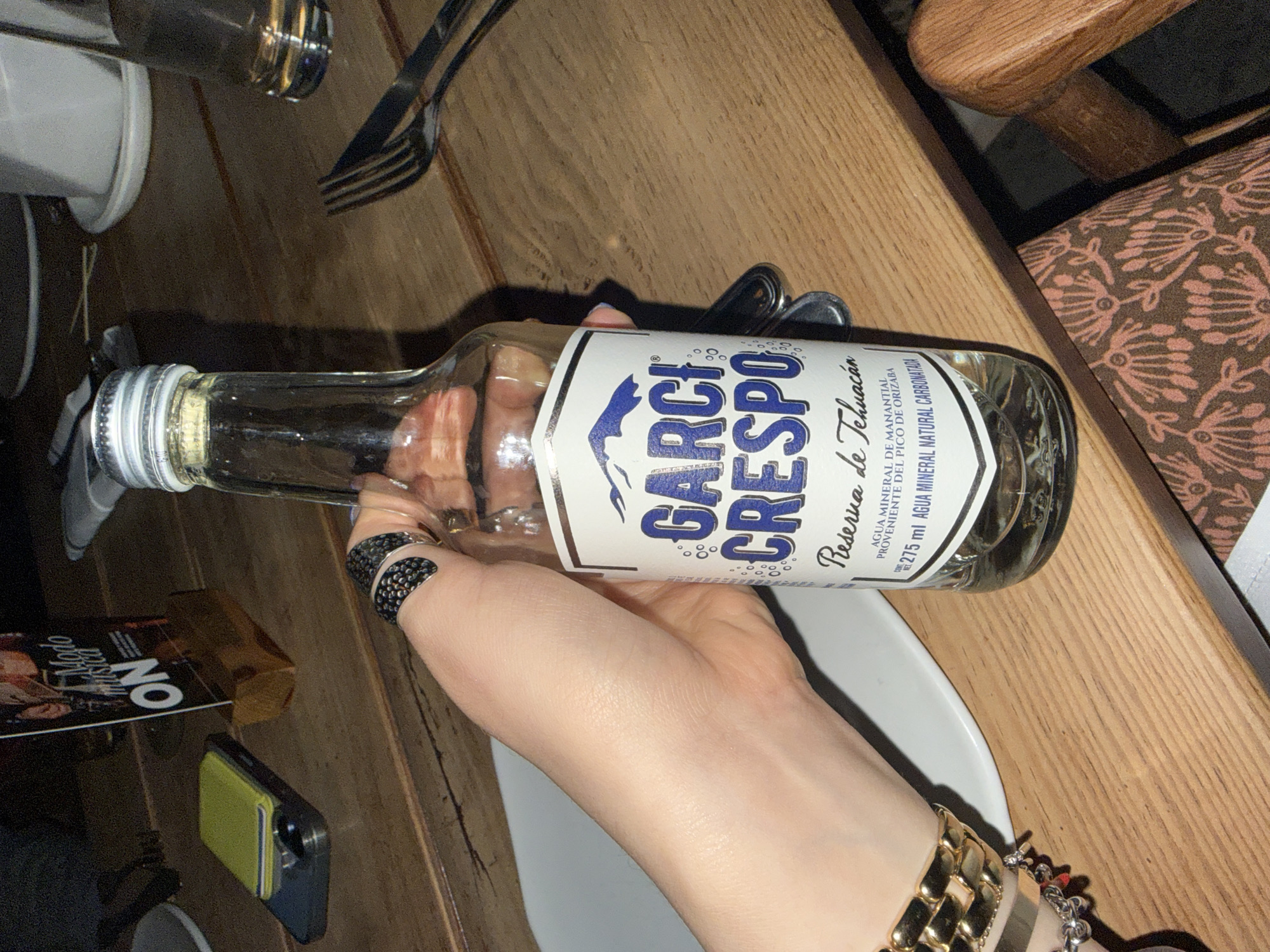 The image shows a hand holding a glass bottle of Garci Crespo mineral water. The bottle is labeled "Reserva de Tehuacán," indicating the water's origin from the Pico de Orizaba region. The hand has several rings and bracelets on it. The bottle is near silverware on a table. A magazine and a phone lie on the wooden surface. The setting appears to be a restaurant, possibly in Mexico City, Mexico, given the product's origin.