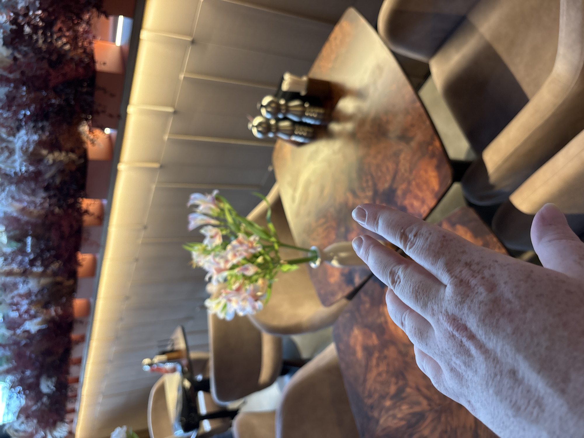 The image shows a close-up view of a hand with freckles and a scar on the back, resting on a table. The table has a polished, wood-grain surface with swirling patterns of brown and orange. In the background, several chairs with upholstered seats and backs are visible, appearing to be in a restaurant or lounge setting. There is a vase with a bouquet of light-colored flowers, possibly lilies or alstroemerias, placed on the table. The lighting suggests an indoor environment, possibly during the day or evening, with warm artificial light reflecting off surfaces. To the left, a blurry section of a wall with decorative elements and vertical light fixtures is visible, hinting at the overall decor. The scene is captured at a slightly downward angle, focusing on the hand and the immediate tabletop. There is no visible text in the image. The overall impression is one of casual observation or a moment captured while seated at a table.