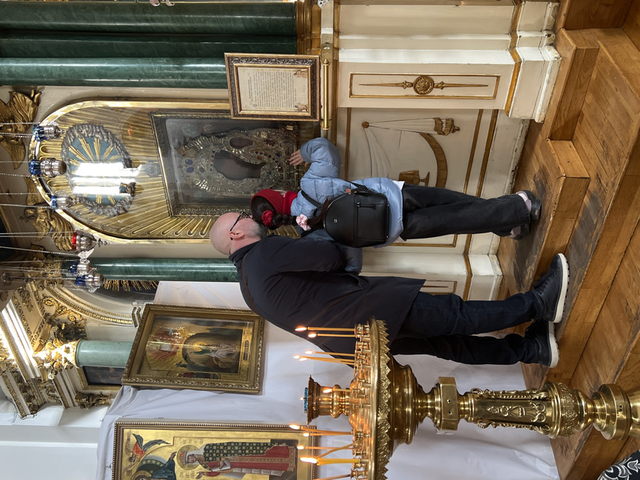 Inside a grand Orthodox cathedral, likely the Kazan Cathedral in Saint Petersburg, Russia, a man and a young girl stand before a prominent, ornately framed icon. The central icon, possibly the Kazan Icon of the Mother of God, is encased behind glass within a large, gilded frame featuring a detailed metal riza adorned with jewels. Above the icon is a golden sunburst motif and several hanging vigil lamps. To the right of the main icon, a framed text panel is displayed.
On the left side of the scene, a large, ornate gilded brass candlestick holds numerous lit yellow candles. Two additional framed icons are visible on the far left, one depicting a standing saint and another a religious scene. The architectural background features green marble columns, gilded decorative elements, and white walls.
The man, bald and bearded with glasses, wears a dark long coat, dark pants, and dark sneakers with white soles. He leans forward, his head close to the girl's, looking towards the icon. The young girl, with dark hair tied in a ponytail with a pink scrunchie and wearing a red knit beanie, a light blue puffer jacket, a black backpack, and dark pants, stands on a wooden step. Her right hand is extended, touching the glass surface protecting the icon. Both figures are positioned reverently before the religious artifact. The flooring consists of wooden planks leading up to the platform where they stand.