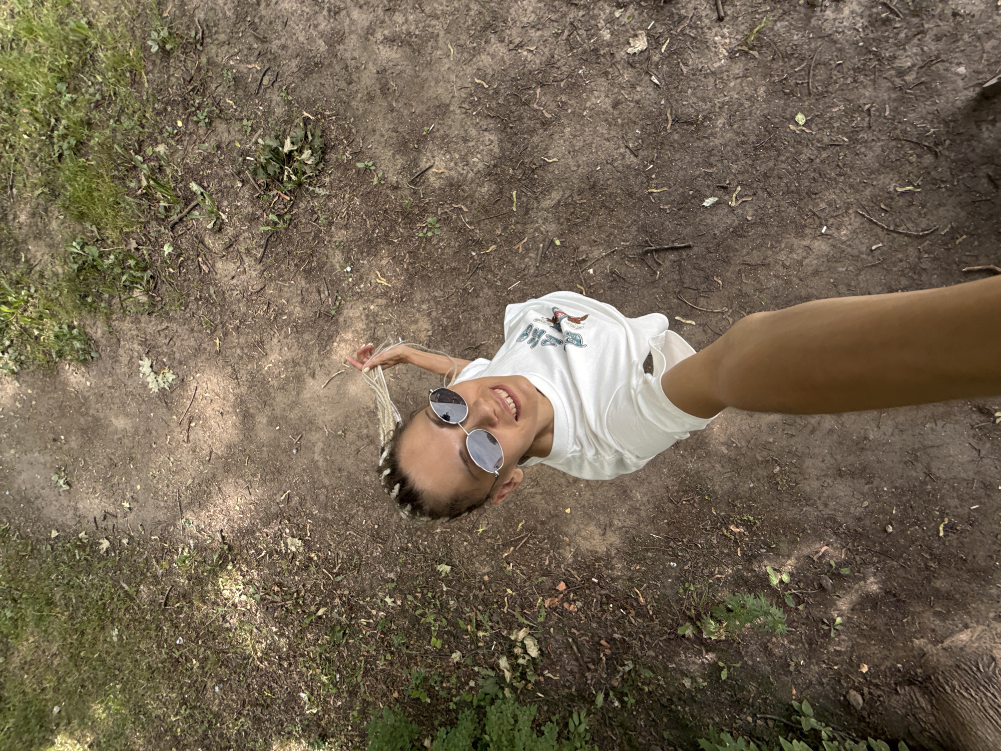 In this overhead, eye-level shot, a young woman is captured from above as she looks up towards the camera with a wide smile. She has light skin and her hair is styled in blonde braids. She is wearing a white t-shirt and silver-rimmed sunglasses. The t-shirt has a graphic design on the front, but the text is illegible.
The woman is standing on a path that is a mix of dirt and grass. The ground is covered in small twigs, leaves, and scattered green foliage. In the background, more grass and dirt are visible. The lighting suggests it is daytime, likely outdoors, with some dappled sunlight filtering through unseen trees. The overall mood is cheerful and candid, indicating a relaxed moment outdoors. The location is not specified, but the environment suggests a natural setting like a park or trail.