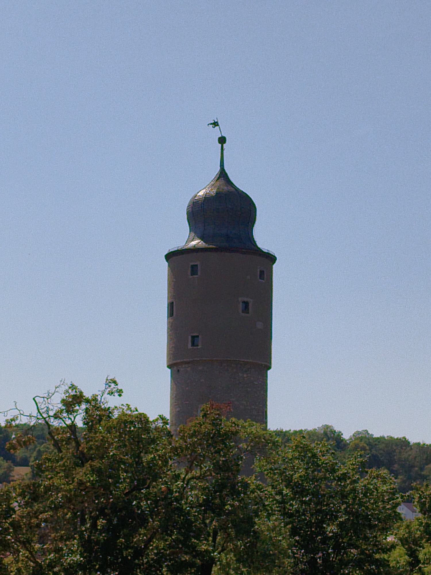 The image shows a tall, cylindrical stone tower with a dark, rounded roof topped with a weathervane. The tower has several small, rectangular windows on its upper section and a stone-like texture on its lower half. In the foreground, a dense canopy of green trees partially obscures the view of the tower. The sky is clear and blue. This is the "Bayersturm" tower, located in Ochsenfurt, Germany.