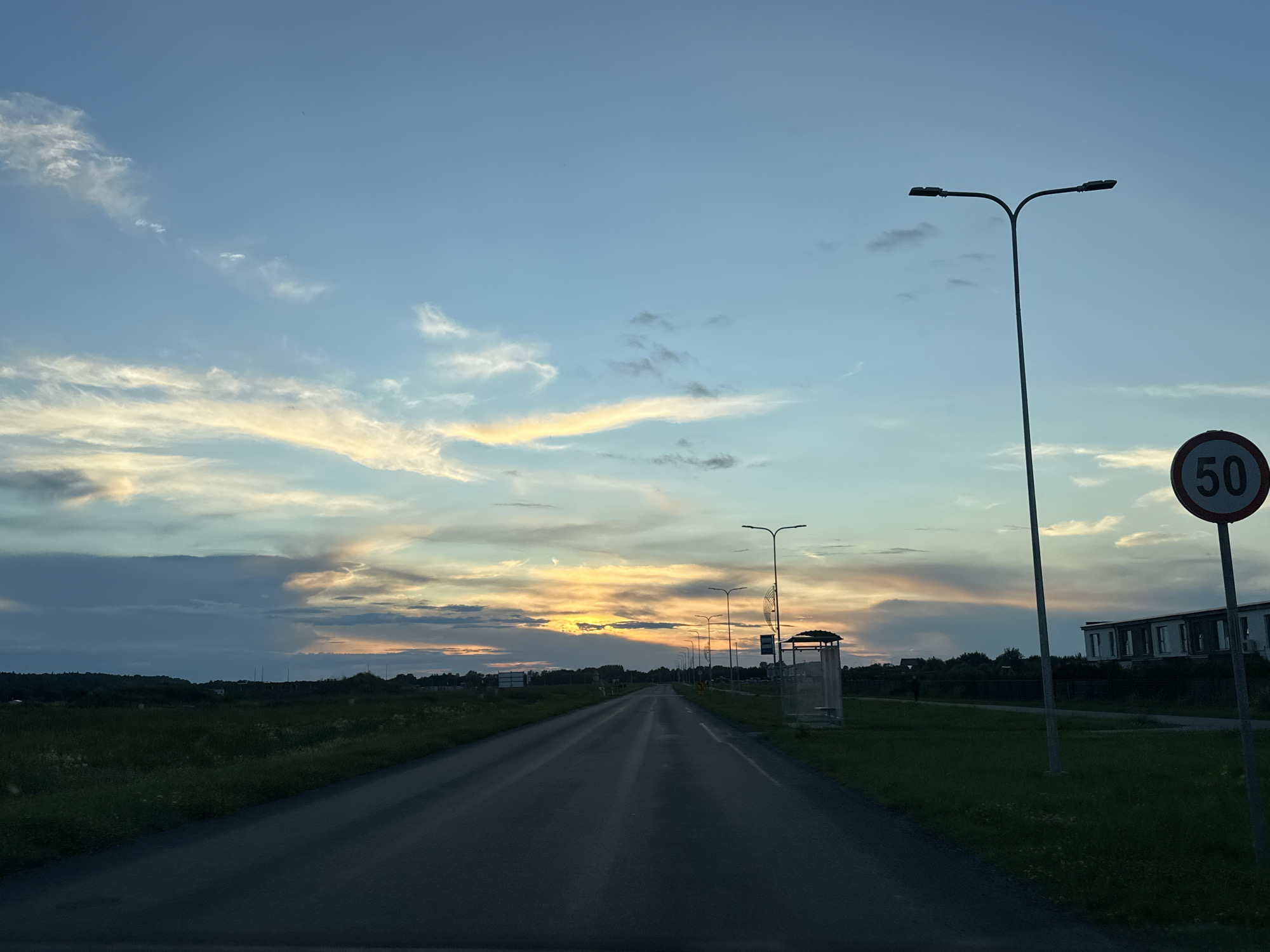 The image depicts a roadside view at dusk in Tiskre, Estonia. A paved road extends into the distance, flanked by green grass and vegetation on either side. Several streetlights line the right side of the road, and a bus stop shelter is visible. Further down the road, a two-story building with a modern facade stands. A circular speed limit sign with the number 50 is prominently displayed on a pole near the road. The sky is a gradient of blues, with streaks of orange and yellow from the setting sun painting the clouds. The overall scene suggests a quiet, suburban or rural atmosphere during twilight.