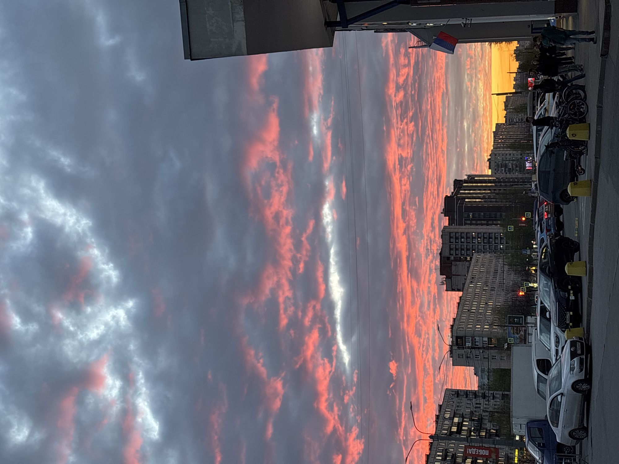 The image captures a vivid sunset over a street in Saint Petersburg, Russia. The sky is filled with striated clouds, predominantly grey but illuminated with streaks of orange and pink from the setting sun. In the distance, a cityscape silhouette features apartment blocks and taller buildings. Along the right edge of the frame, there's a row of parked cars of varying makes and models and a small group of people standing on the sidewalk. A Serbian flag is visible, attached to a building on the street. Overhead electrical wires extend across the scene.