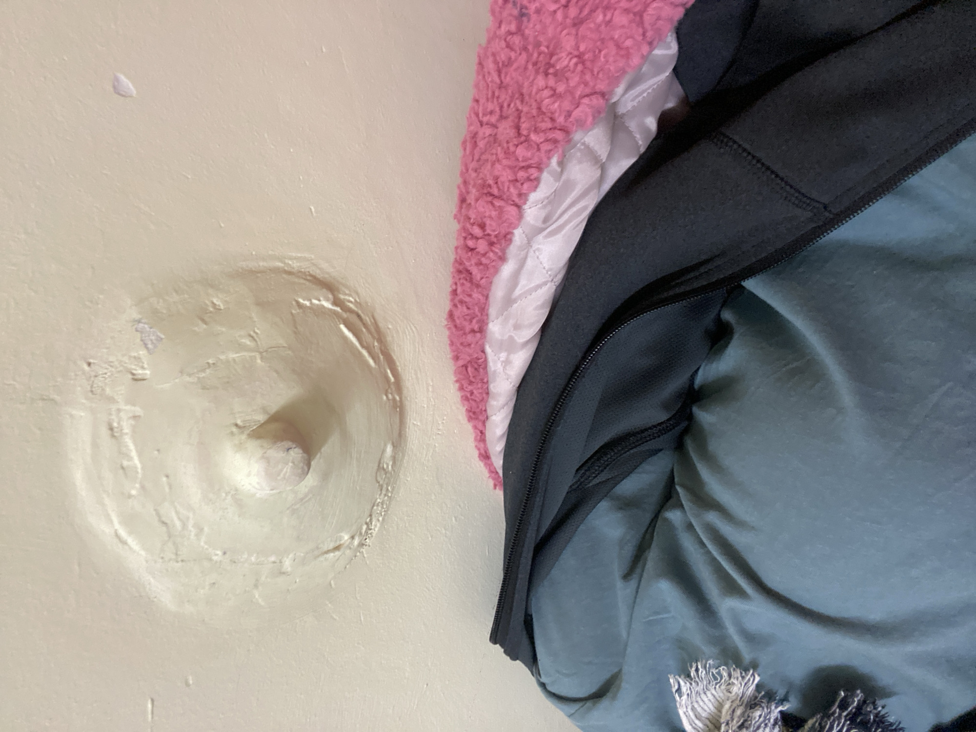 The image displays a close-up, slightly angled view of clothes hanging on a wall-mounted fixture. On the left side, a light, possibly off-white or cream-colored wall features a prominent, circular indentation or buildup around a central white peg or hook. This area exhibits a rough, uneven texture, suggesting a patched or poorly finished wall repair.
To the right, several layers of clothing are visible, draped and folded. The top layer is a vibrant pink, fluffy fabric, likely a faux fur or sherpa material. Beneath it, a white or very light pink quilted fabric with a diamond stitch pattern can be seen. Further down, a dark grey or black garment, possibly a jacket or hoodie, is visible, featuring a black zipper running vertically. The lowest visible layer of clothing is a teal or dark blue-green fabric, appearing soft and somewhat wrinkled. At the bottom right corner, the fringes of what appears to be a scarf or blanket, with threads in white, grey, and dark blue, are partially visible.
The scene is an indoor setting, likely a closet or room, with soft, ambient lighting. No people are present, and no text is visible within the frame.