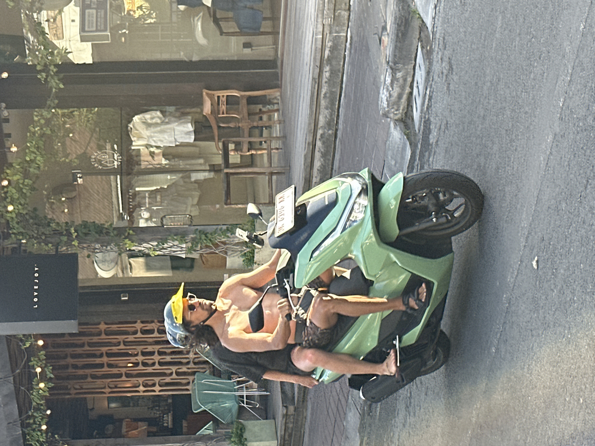 In North Kuta, Indonesia, two individuals are riding a light green scooter on a paved street. The person in the front, driving the scooter, is a bare-chested male wearing a black helmet with a yellow visor and sunglasses. He has long, dark hair and is wearing patterned shorts and dark sandals. The second person, a passenger, is seated behind him and is also wearing a helmet. This passenger is wearing a dark shirt and has their legs visible. The scooter has a white license plate with text that is partially obscured. The background features a building with a black sign displaying "LOVEJOY" and an entrance with decorative wooden latticework. The building's facade includes large glass windows, through which clothing items and mannequins can be seen displayed in a store. Vines with small lights are draped over the storefront. To the left of the scooter, a light blue chair is visible on a paved area. The street surface is asphalt with some cracks and a concrete curb. The lighting suggests it is daytime, with sunlight casting shadows.