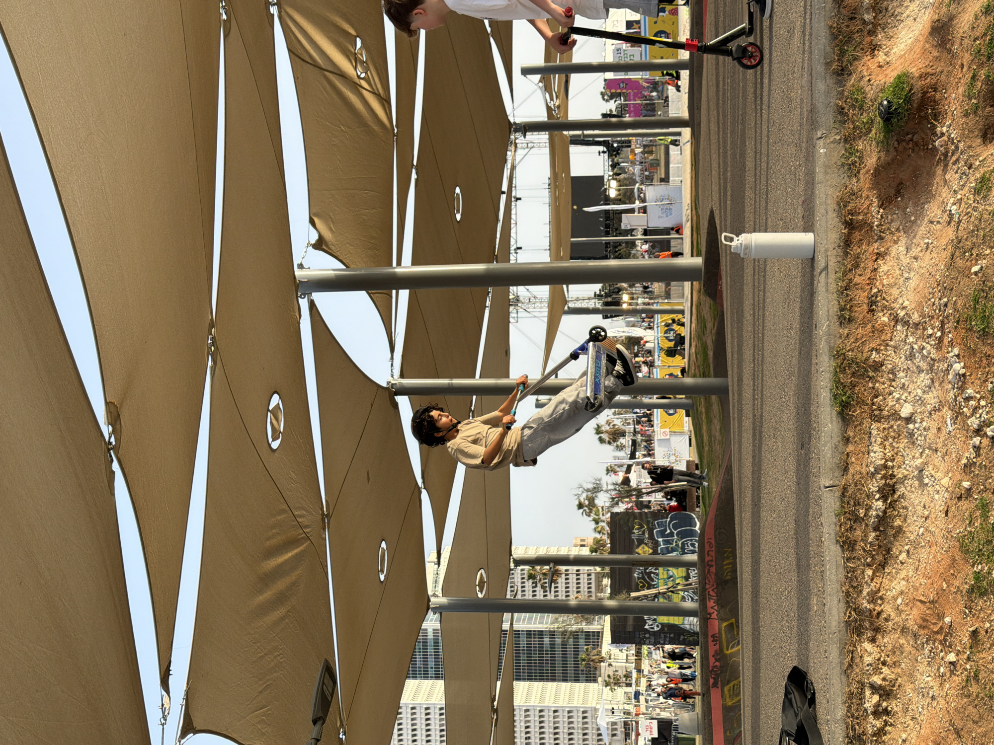 A young man performs tricks on a scooter under a shaded area in Tel Aviv, Israel. The shaded area is composed of multiple triangular, tan-colored awnings with circular cutouts, supported by gray metal poles. In the background, there are buildings and what appears to be a market or public event. The man is wearing a helmet, a tan t-shirt, and light gray pants. There is a pedestrian walkway next to the man. On the other side of the walkway, there is a patch of dirt and some vegetation. A water bottle sits nearby on the walkway.