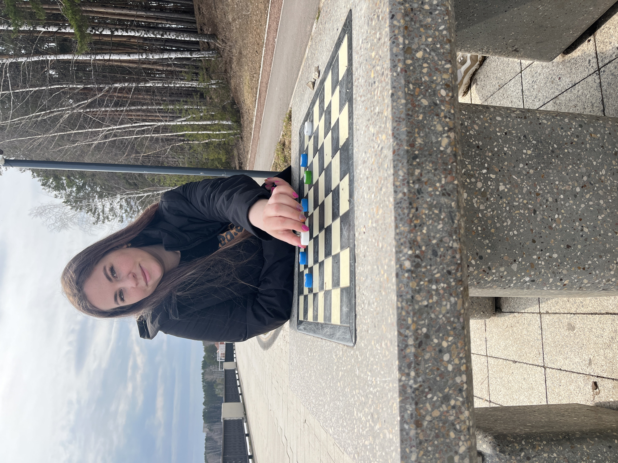 A young woman with long brown hair wearing a black jacket is captured at a park, positioned at a concrete chess table in Chaikovsky, Russia. She is in the process of moving a chess piece, specifically a white piece, on the chess table. The chess table itself is constructed from a concrete material with a speckled pattern. Behind the chess table, a paved pathway runs alongside a wooded area featuring trees with sparse foliage. In the distance, a body of water can be seen, along with building infrastructure along the water's edge, under a sky with patchy clouds. The scene is generally bathed in daylight, indicating daytime conditions.