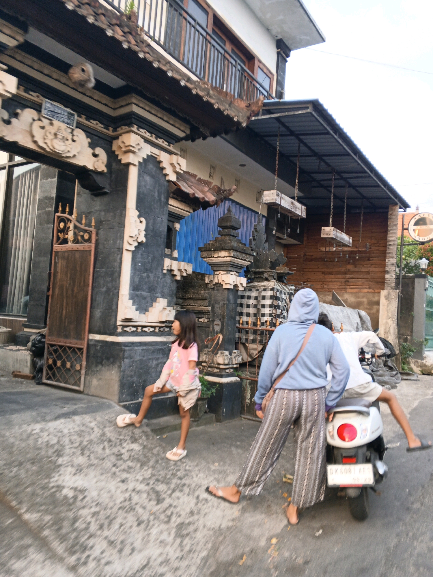 In Denpasar, Indonesia, a young girl in a pink t-shirt and shorts stands in the foreground on a paved surface. She is facing right, with her left leg bent and extended forward. Another person, wearing a blue hooded sweater and striped pants, is positioned behind her and to the right, straddling a white scooter. The scooter's rear is facing the viewer, with a red taillight and a license plate clearly visible. Behind the scooter, another individual is seated. The background features an outdoor environment with a building showcasing traditional Balinese architecture, including carved stone elements and a wooden door. A metal awning extends from the building, supported by chains. The overall scene depicts a street or an alleyway in a residential or commercial area.