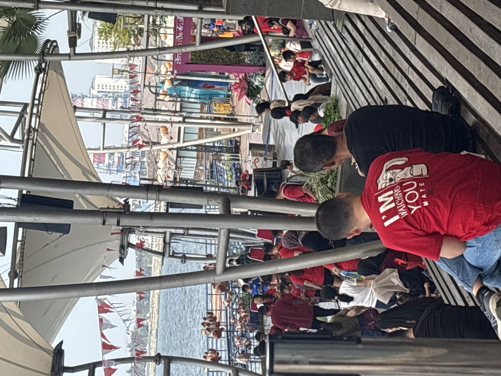 The image shows a crowd of people gathered on an outdoor promenade, likely in North Jakarta, Indonesia, given the presence of industrial structures and waterfront. The scene is characterized by a series of slanted wooden benches where many individuals are seated, facing various directions. In the foreground, a man in a red t-shirt with a graphic design and text that reads "I'M WATCHING YOU MATE" is prominently featured. Beside him, another man in a black t-shirt is seated. Further back, numerous other individuals, dressed in casual attire, are scattered throughout the area. Some appear to be standing, while others are seated on the benches or on the ground. In the background, there is a body of water, and beyond that, a skyline with buildings and industrial facilities, including tall towers with red and white striped elements. Decorative elements like red and white triangular flags and hanging lanterns are visible, suggesting a festive or event-like atmosphere. The overall setting suggests a public gathering space near a waterfront in an urban environment.
