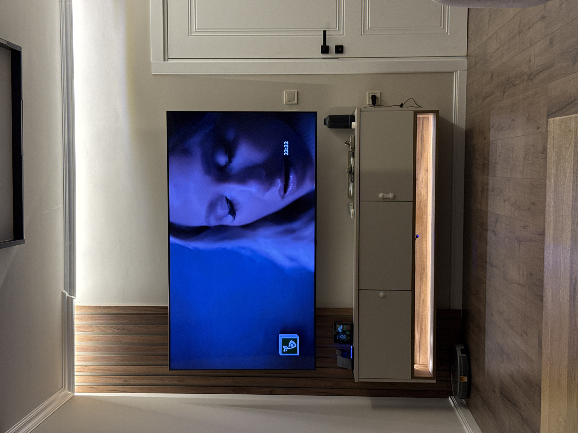 The image captures an indoor living space, featuring a large television screen displaying a woman's face with closed eyes, bathed in a blue light. The digital time "23:22" is visible on the screen, indicating a late hour. Below the screen, a small illuminated icon, possibly an application logo, is displayed.
To the right of the television, a modern, light-grey cabinet unit stands vertically, featuring multiple closed compartments and a central illuminated section with warm lighting, revealing vertical wooden slats within. Above this cabinet, a black cylindrical device is mounted on the wall, possibly a smart speaker or camera. Various electrical outlets, plugs, and cables are visible around the cabinet and above the TV.
The lower portion of the wall behind the TV and cabinet is adorned with horizontal wooden slatted paneling. Above this paneling, the wall is painted a light, neutral color. At the top of the image, a white, paneled door or built-in cabinet with decorative molding and dark handles or fixtures is partially visible. The room features light wood-patterned flooring on the right side and a light-colored baseboard along the bottom. On the far left, a white wall section with decorative trim is present. A small, illuminated device is visible on the floor next to the cabinet, possibly a charging dock for a robot vacuum. The overall ambiance suggests a calm, modern interior setting in Plovdiv, Bulgaria, during the late evening.