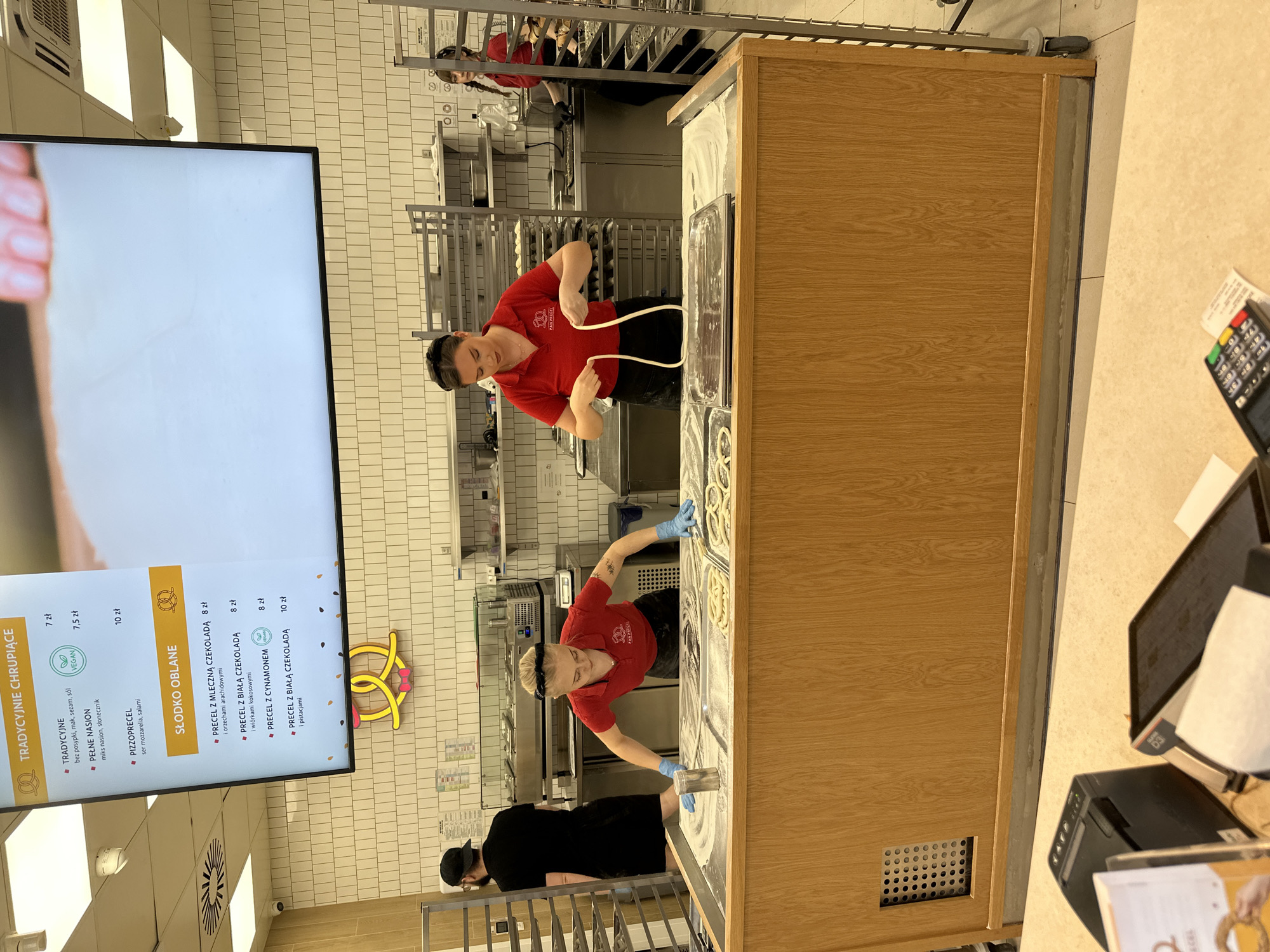 The image shows the interior of a "PAN PRECEL" bakery, which is a franchise specializing in pretzels. Two employees wearing red shirts with the "PAN PRECEL" logo are working at a wooden countertop, preparing pretzels. One employee is stretching dough, and the other appears to be shaping pretzels. They are both wearing gloves. Behind them, there are stainless steel racks with baking trays. There is also a partially visible third person in black near the front of the counter, who may be another employee. On the left, there is a digital menu board displaying pretzel varieties and prices in Polish zlotys (zł). The menu includes "TRADYCYJNE CHRUPIĄCE" (traditional crispy) and "SŁODKO OBLANE" (sweet coated) options. A point-of-sale system is visible in the foreground. The location is Wrocław, Poland, based on the menu and the name of the bakery, "PAN PRECEL" which is a local business. The wall has white tiled backsplash.