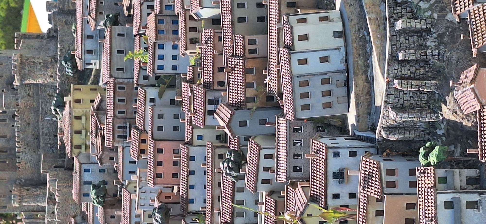 This is a detailed miniature model of a Mediterranean village with a castle in the background, likely situated in Rimini, Italy. The model showcases a dense collection of small, multi-story houses with red tiled roofs, clustered closely together on a hillside. The architecture suggests a historical European town, with pastel-colored facades in shades of yellow, blue, pink, and white, punctuated by numerous dark square windows and some balconies. Small green trees and shrubs are interspersed among the buildings, and a prominent stone castle with turrets stands at the top of the scene, overlooking the village. The stonework of the castle and the retaining walls supporting the village suggests an ancient or medieval setting. The overall impression is of a well-crafted diorama designed to represent a picturesque Italian coastal town.