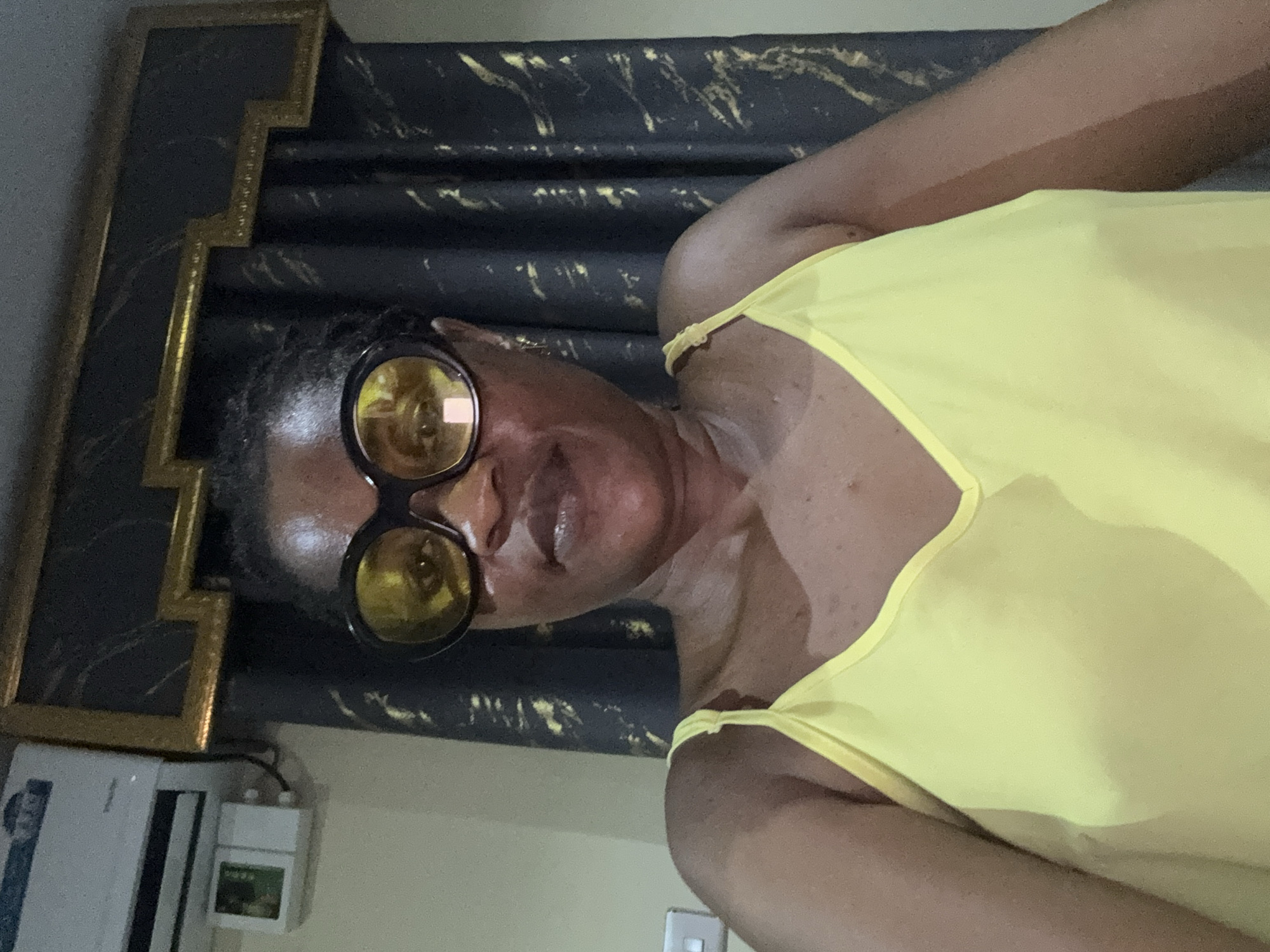 The image shows a woman wearing a yellow tank top and large round sunglasses with yellow lenses. She is positioned in front of a dark, textured curtain with gold accents. A decorative gold-trimmed frame is visible to the left of the woman, partially obscuring a white air conditioning unit and a small white box with green text on it. The woman's skin appears slightly shiny, possibly from sweat, and her expression is neutral. The lighting suggests it is daytime. The scene is indoors, with the air conditioning unit indicating a possibly warm climate. The woman is looking towards the camera.