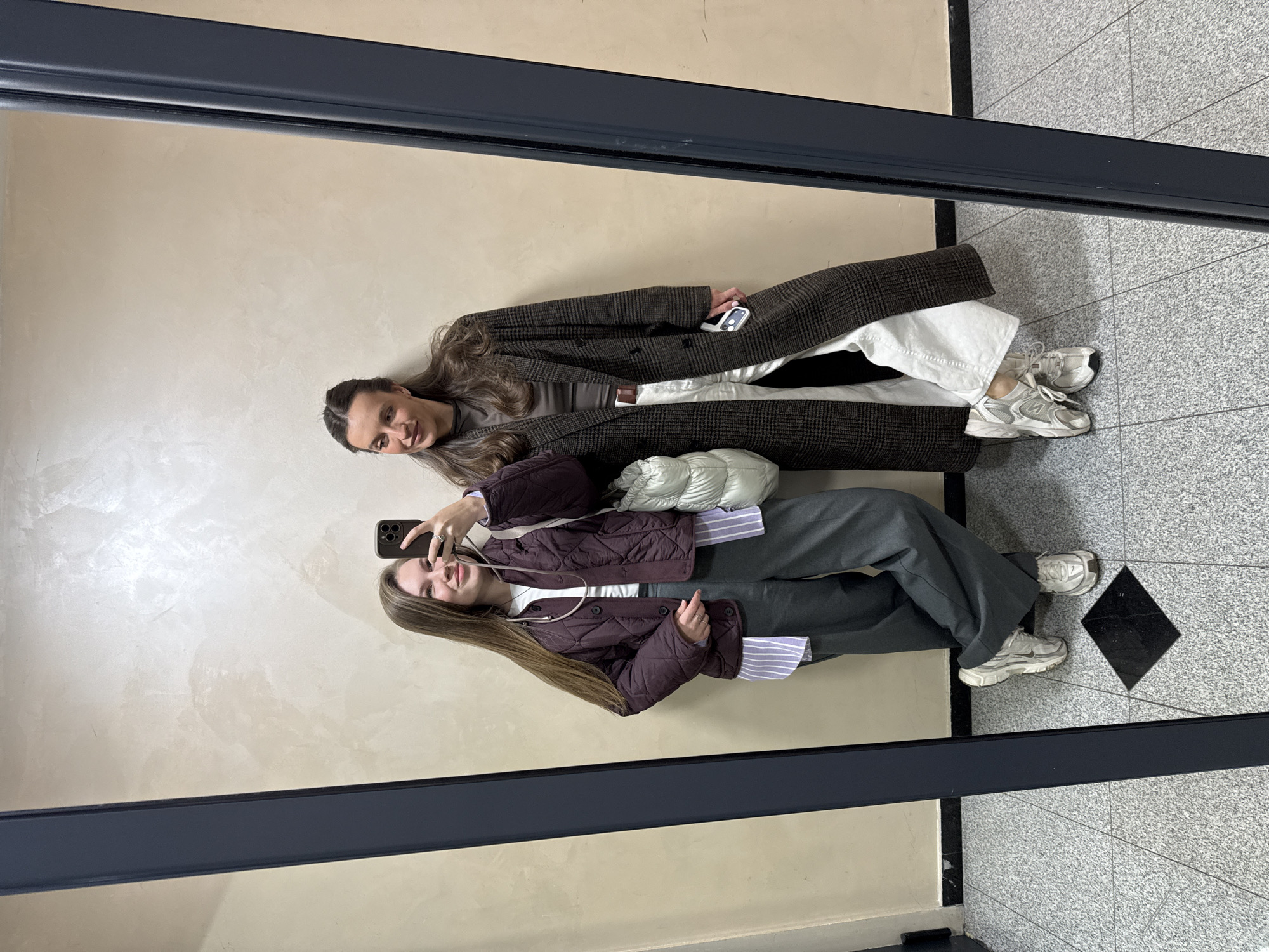Two women are reflected in a mirror. The mirror is framed by a dark gray or black frame. The walls behind the women are beige. The floor is gray with a repeating pattern, including dark diamond shapes.
The woman in the foreground reflection is holding a smartphone and taking a selfie. She has long brown hair and wears a short quilted burgundy jacket over a striped shirt, gray pants, and white sneakers. A white bag is slung over her shoulder.
The woman in the background reflection has long brown hair. She is wearing a brown coat over a brown shirt, white pants, and white sneakers. She is holding a white object in her hand.
The scene appears to be indoors, possibly in a public space. There is no specific information to identify the exact location within Vozdovac Urban Municipality, Serbia.