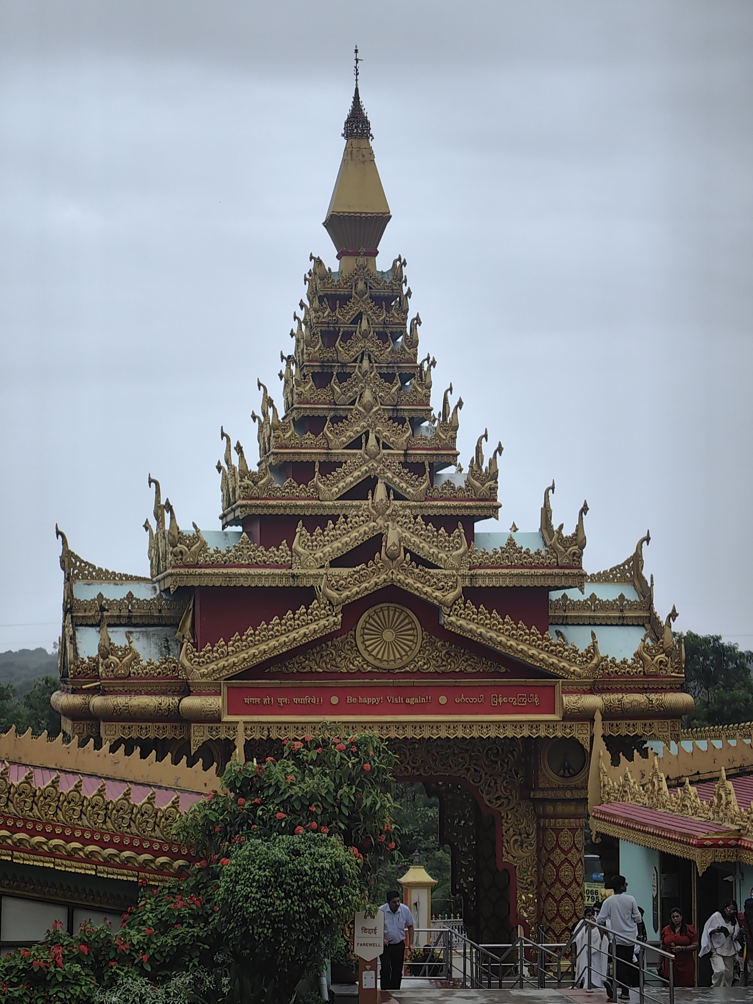 This is a vertical, eye-level, full shot of the entrance to the Global Vipassana Pagoda, located in Mumbai, India. The structure is elaborately decorated in gold and red, with multiple tiers and ornate architectural details. The sky is overcast. The foreground features lush green trees with red flowers, and a pathway leading towards the entrance. Several people are visible in the image, some walking towards the entrance and others near it. One person in the foreground is wearing a light blue shirt and dark pants, while others are in white or red clothing. A sign with the word "FAREWELL" and its Hindi equivalent is visible on the right. The overall scene depicts a serene and culturally significant site.