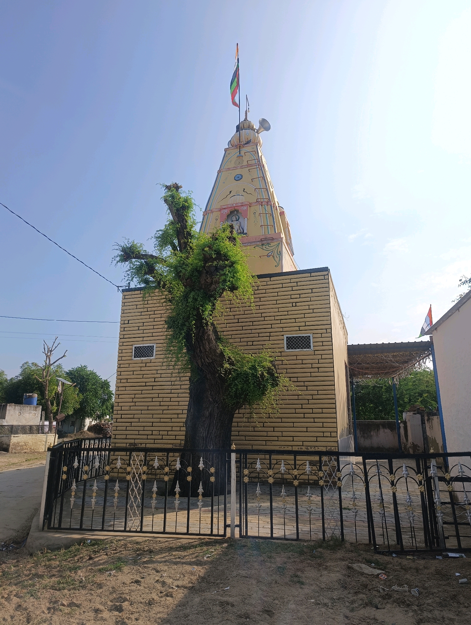 The image shows a religious structure, likely a temple, in Chhapri Bari, India. The temple has a distinctive yellow, conical spire topped with a flag and a speaker. The lower part of the building is constructed with yellow bricks, and a large, leafy tree stands prominently in front of it, obscuring part of the facade. A black metal fence with decorative elements surrounds the area in front of the temple. The environment appears to be a rural or semi-urban setting with some trees and buildings visible in the background under a clear blue sky. The lighting suggests it is daytime. There are no people visible in the image.