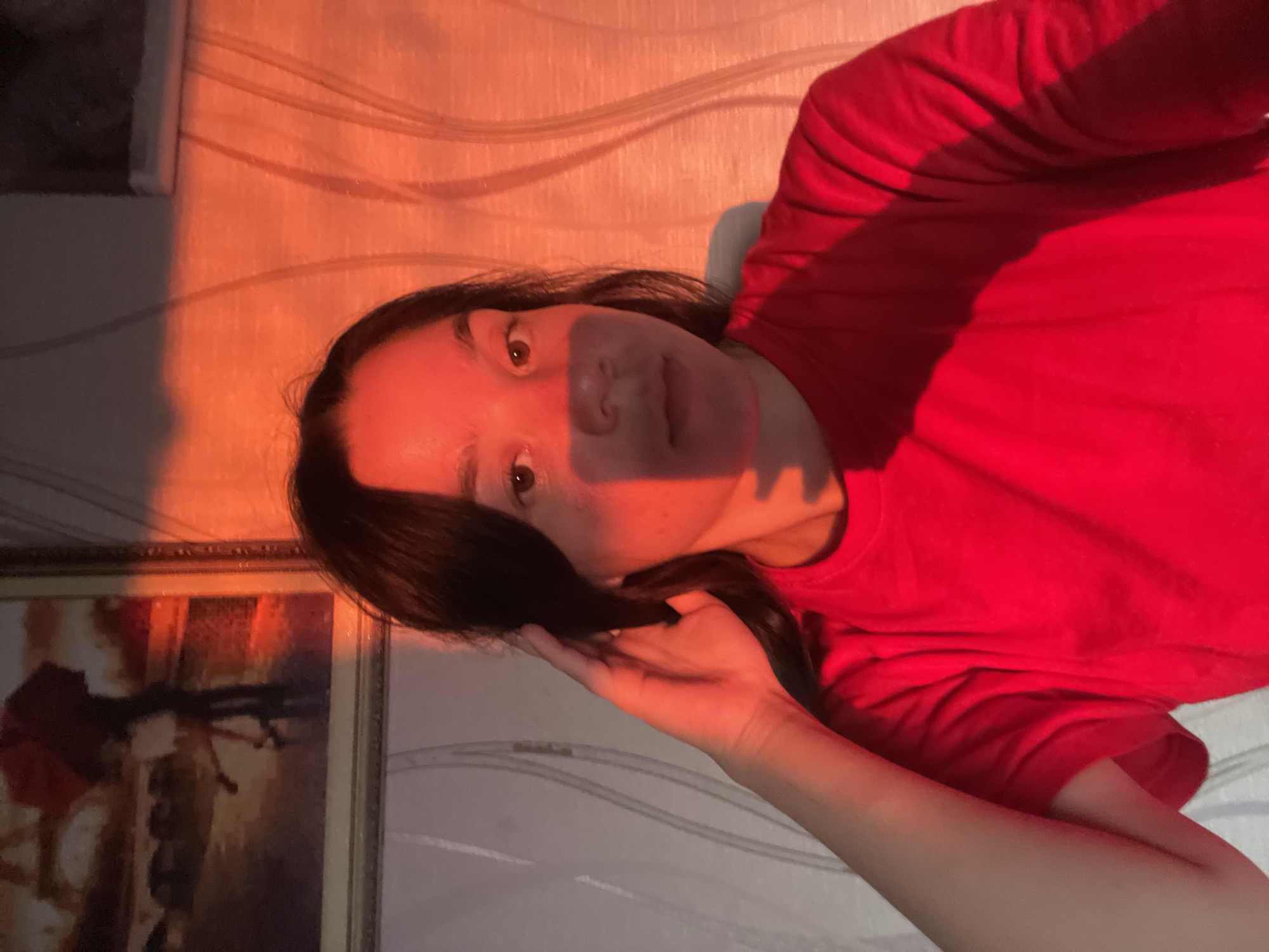 A young person with dark hair is seen from the chest up, looking directly at the camera. They are wearing a red t-shirt and have their left hand raised, touching their hair. The scene is illuminated by a strong red light source, casting dramatic shadows across their face and the background. The background appears to be an interior setting with patterned wallpaper and a framed picture on the wall. The picture on the wall depicts a scene with a red umbrella and two figures, possibly in a city setting. The lighting suggests it might be sunset or a similarly colored artificial light. There is no discernible location in the city of Olkhovka, Russia.