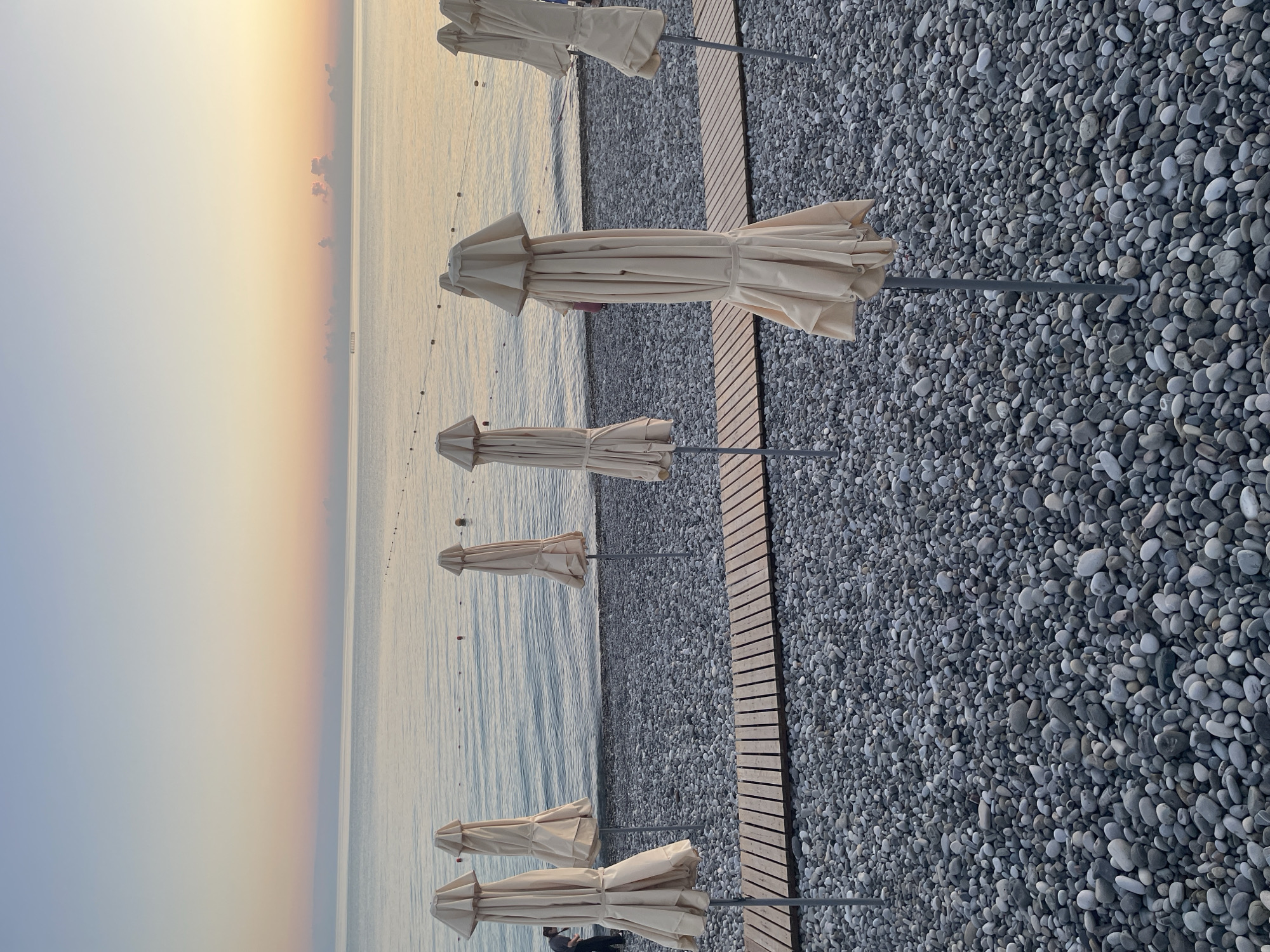 A vertical, eye-level, medium shot captures a pebble beach with folded beach umbrellas lined up. The scene is set at dusk with a tranquil sea and a gradient sky of pale blue, pink, and yellow. The umbrellas are cream-colored and closed, each supported by a grey pole. A wooden pathway made of planks runs horizontally across the lower portion of the frame, interrupting the pebble-covered ground. In the far left background, a person in a dark shirt is partially visible, standing near the shoreline. The sea is calm with faint ripples, and a thin line of buoys stretches across its surface. The overall atmosphere is serene and still.