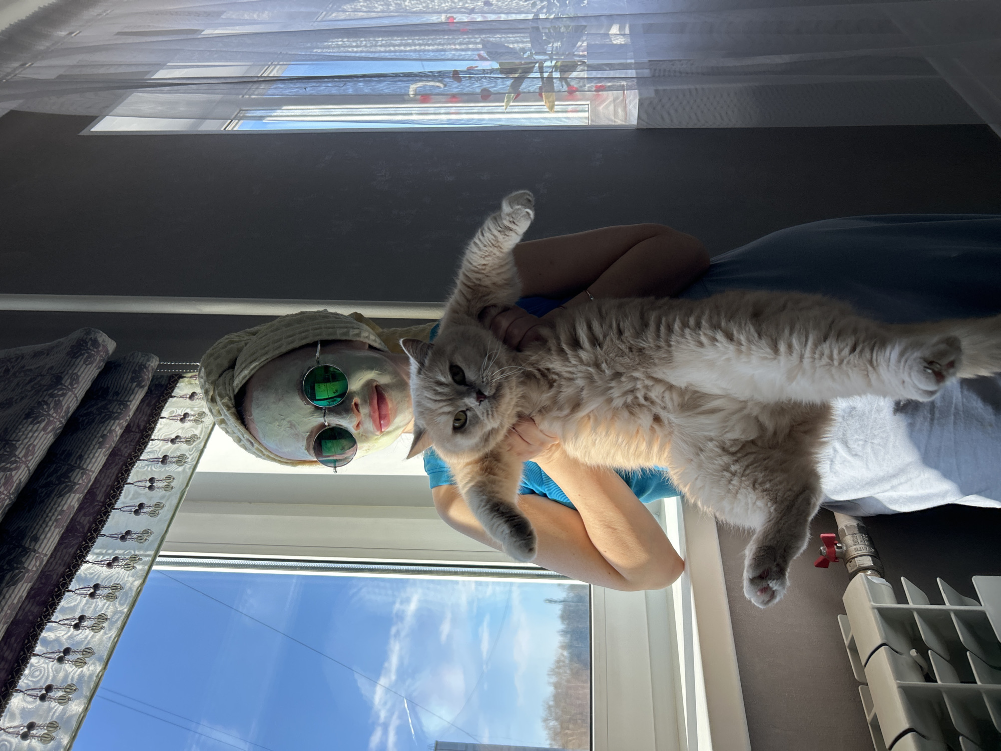 A person wearing a face mask and sunglasses, with a towel wrapped around their head, holds a long-haired cat in front of a window. The person's clothing is partially visible, appearing to be a blue top. Outside the window, a clear blue sky with a few clouds is visible. A radiator is located near the window. The location is Chaikovsky, Russia.