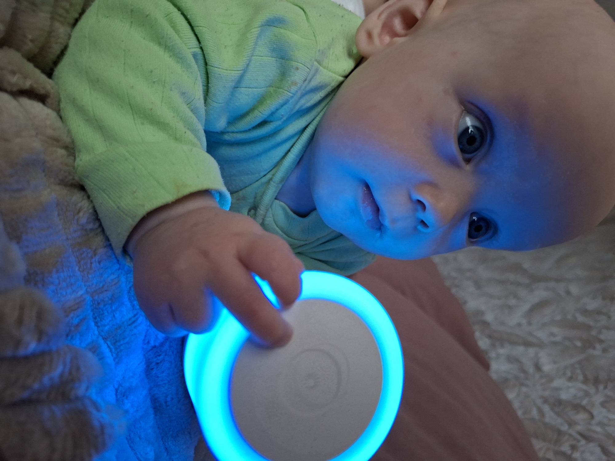 An infant with light skin and blue eyes is depicted indoors, looking intently towards a circular, disc-shaped object held in their right hand. The object emits a prominent blue light from its outer ring, illuminating the infant's face, neck, and upper chest with a blue hue. The infant's right index finger is positioned on the central, light-colored surface of the disc, which bears a subtle, embossed pattern resembling a sun or star. The infant is wearing a long-sleeved, light green garment, visible on the left arm and chest, displaying minor pilling and faint smudges. The infant is positioned on a textured, light brown blanket, which also reflects the blue light. In the background, a light-colored, textured surface, possibly a pillow or wall covering, is visible behind the infant's head, with a segment of a muted pink/peach fabric object situated to the right. The overall lighting conditions are dim, with the blue light source being dominant. The location is Unknown, Unknown.